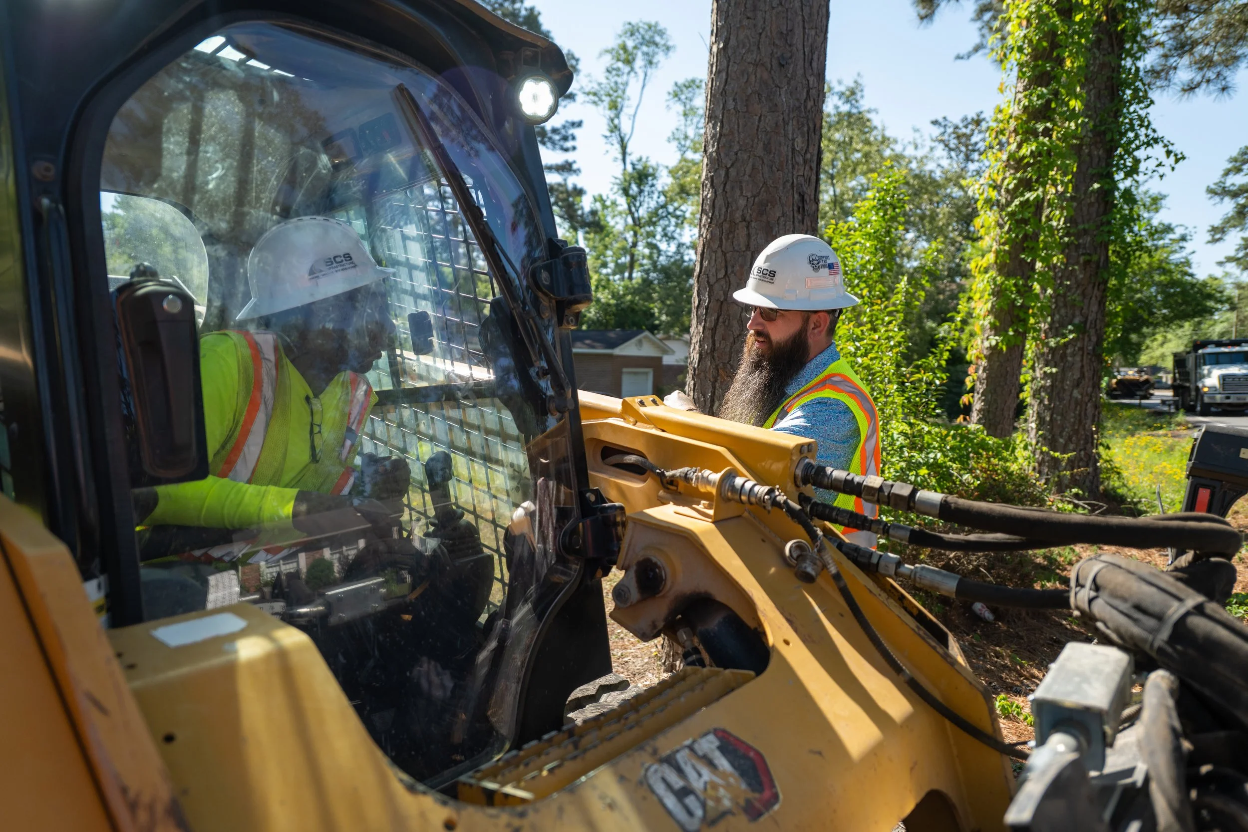 Construction workers wearing helmets and safety gear operating heavy machinery outdoors, with trees and a clear sky in the background.