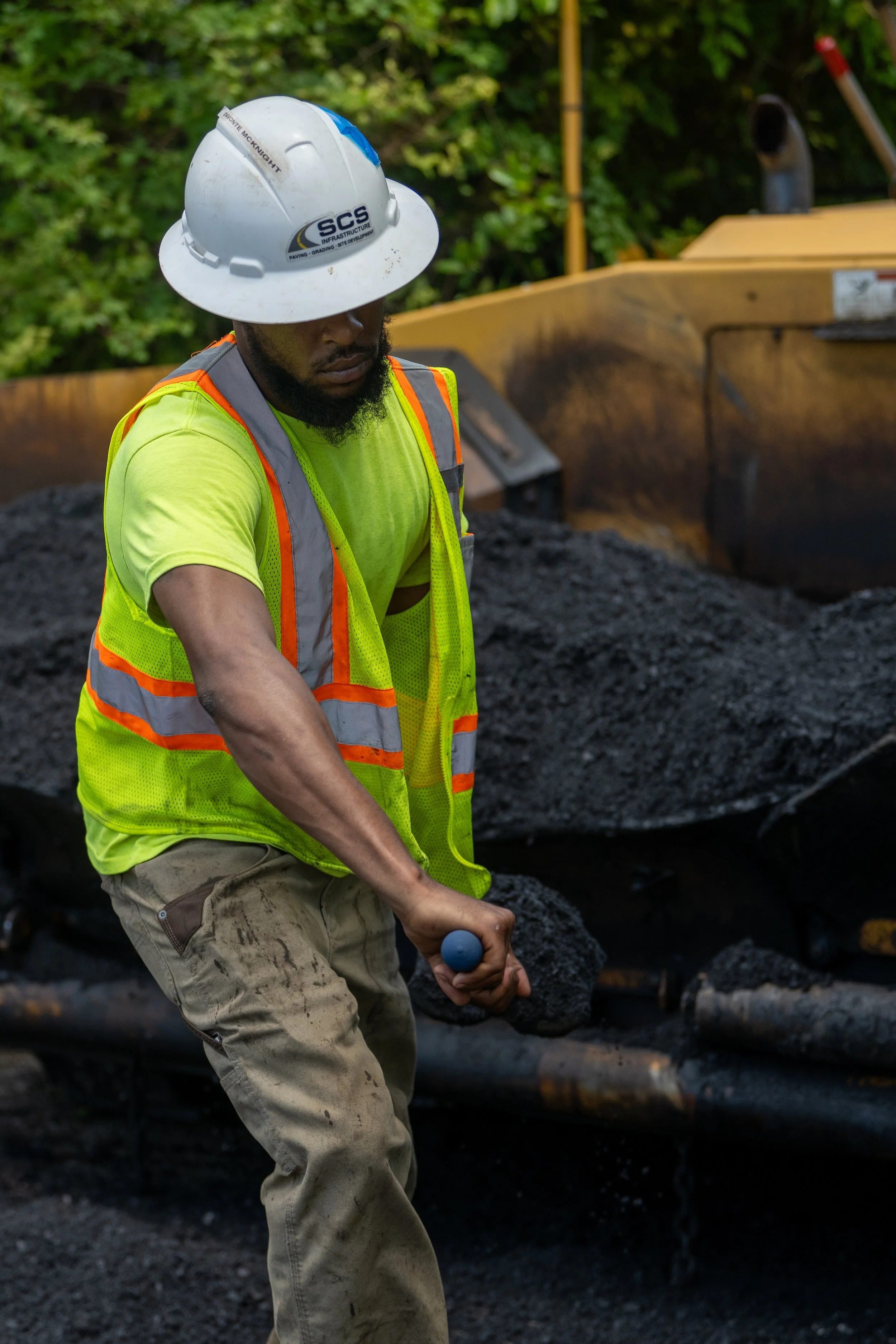 A construction worker wearing a white hard hat, a neon yellow safety vest with orange reflective stripes, and beige work pants, holding a sledgehammer, standing in front of a pile of asphalt and an excavator with a yellow body and black exhaust pipe, with green trees in the background.