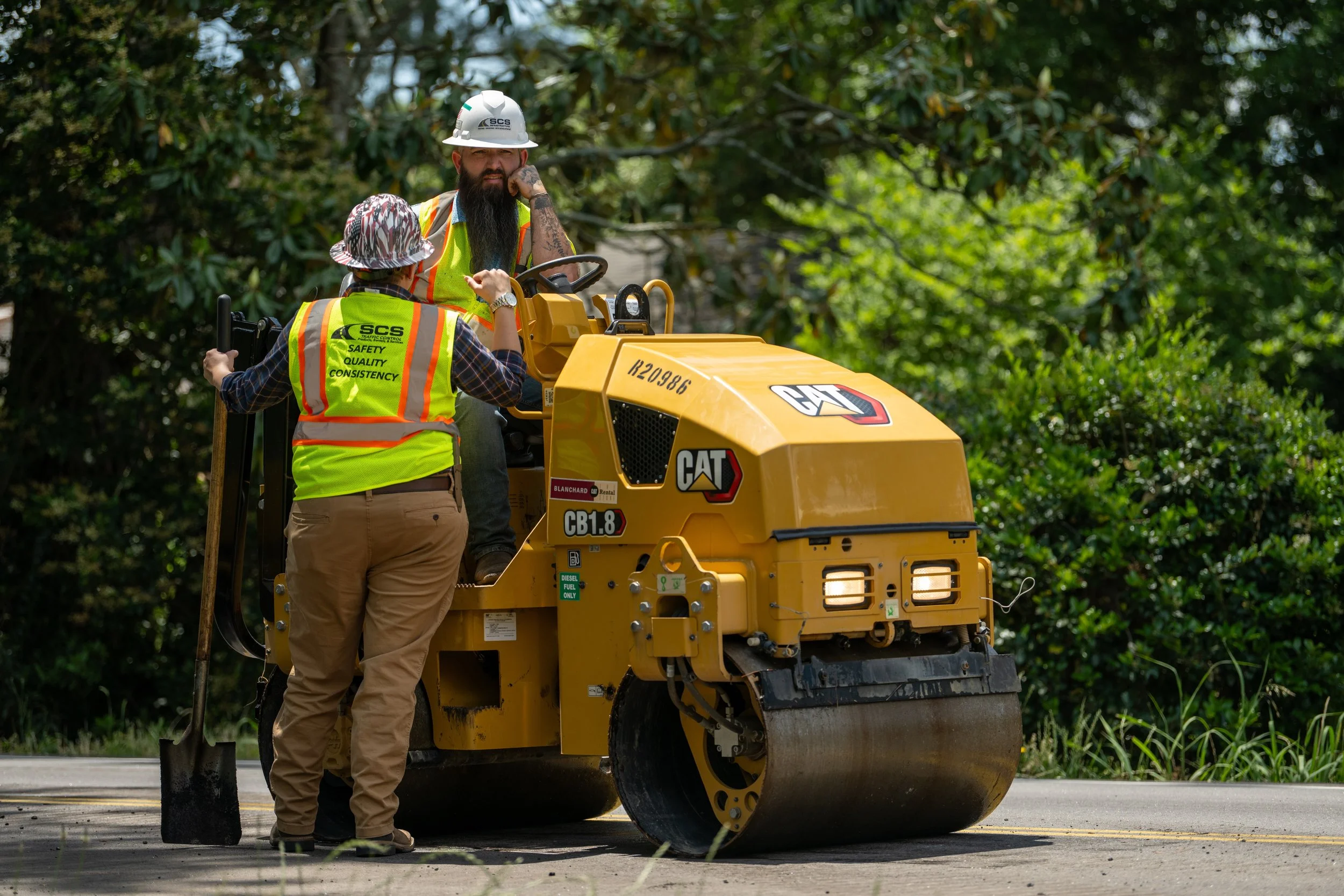 Two workers operating a yellow road roller compactor near a tree on a paved surface during daytime. They are wearing high-visibility safety vests and helmets.