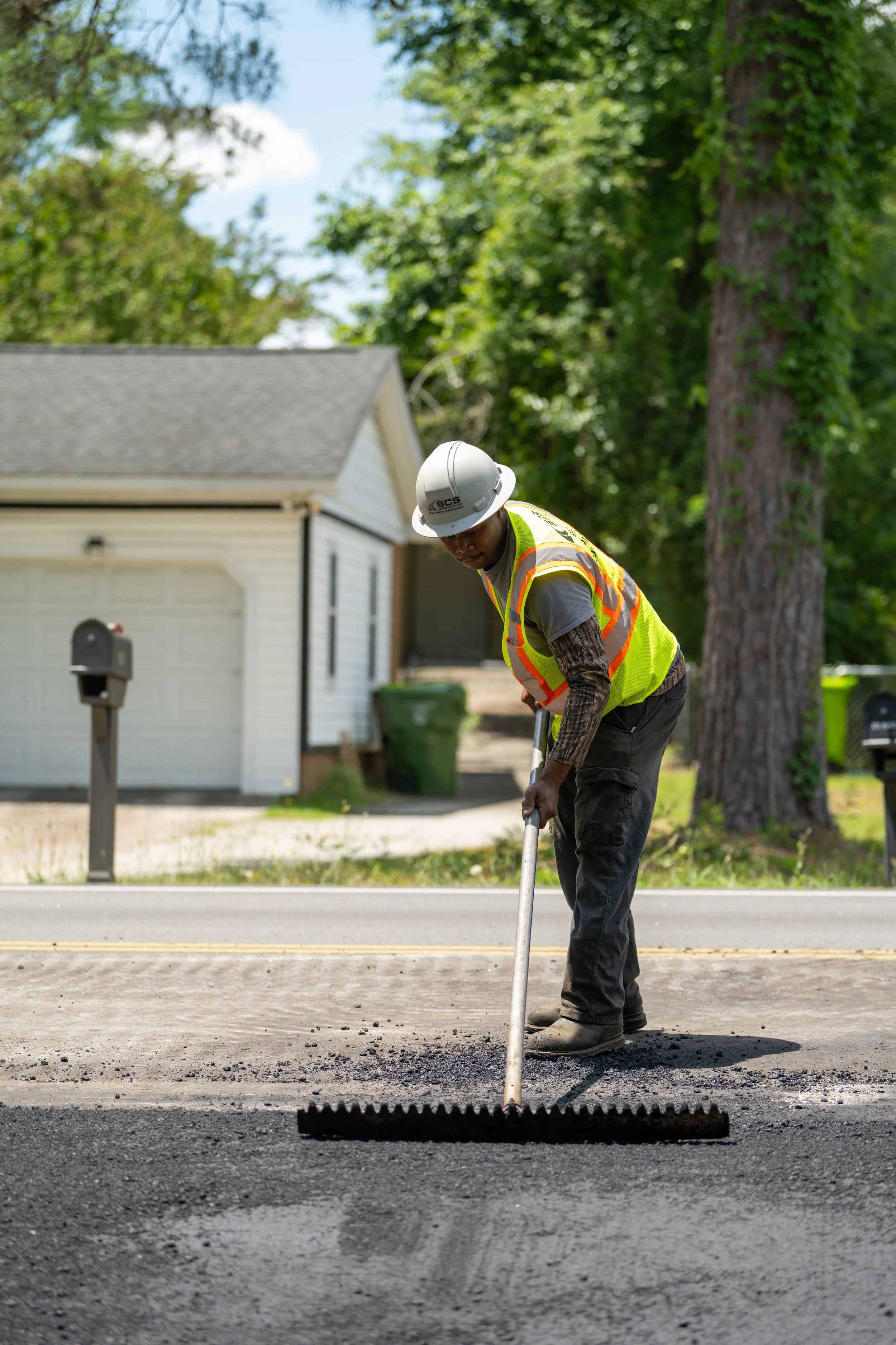 Worker in a yellow safety vest and helmet smoothing fresh asphalt on a residential street.