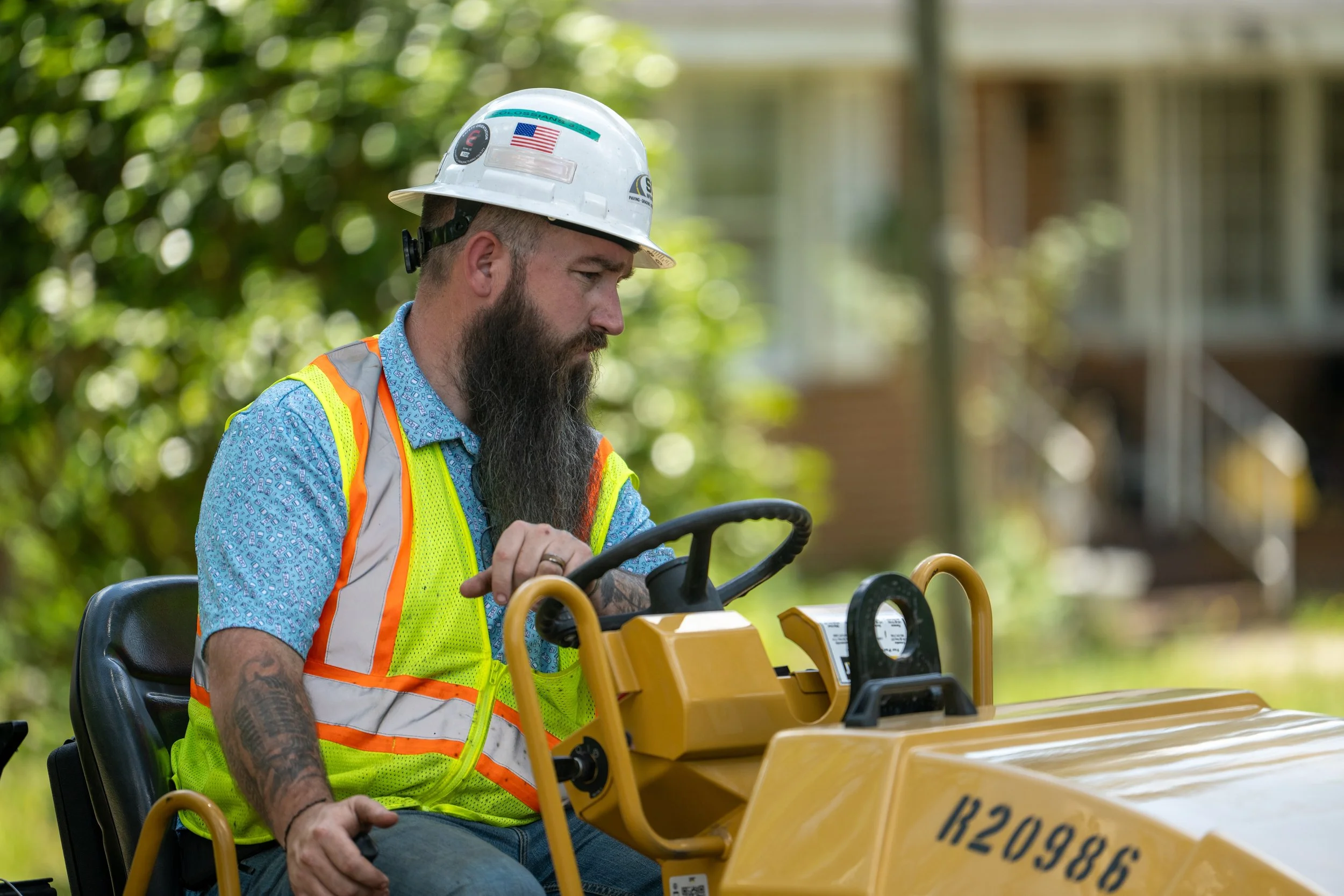 A man with a beard wearing a white hard hat, colorful safety vest, and patterned shirt, operating a yellow construction vehicle outdoors.