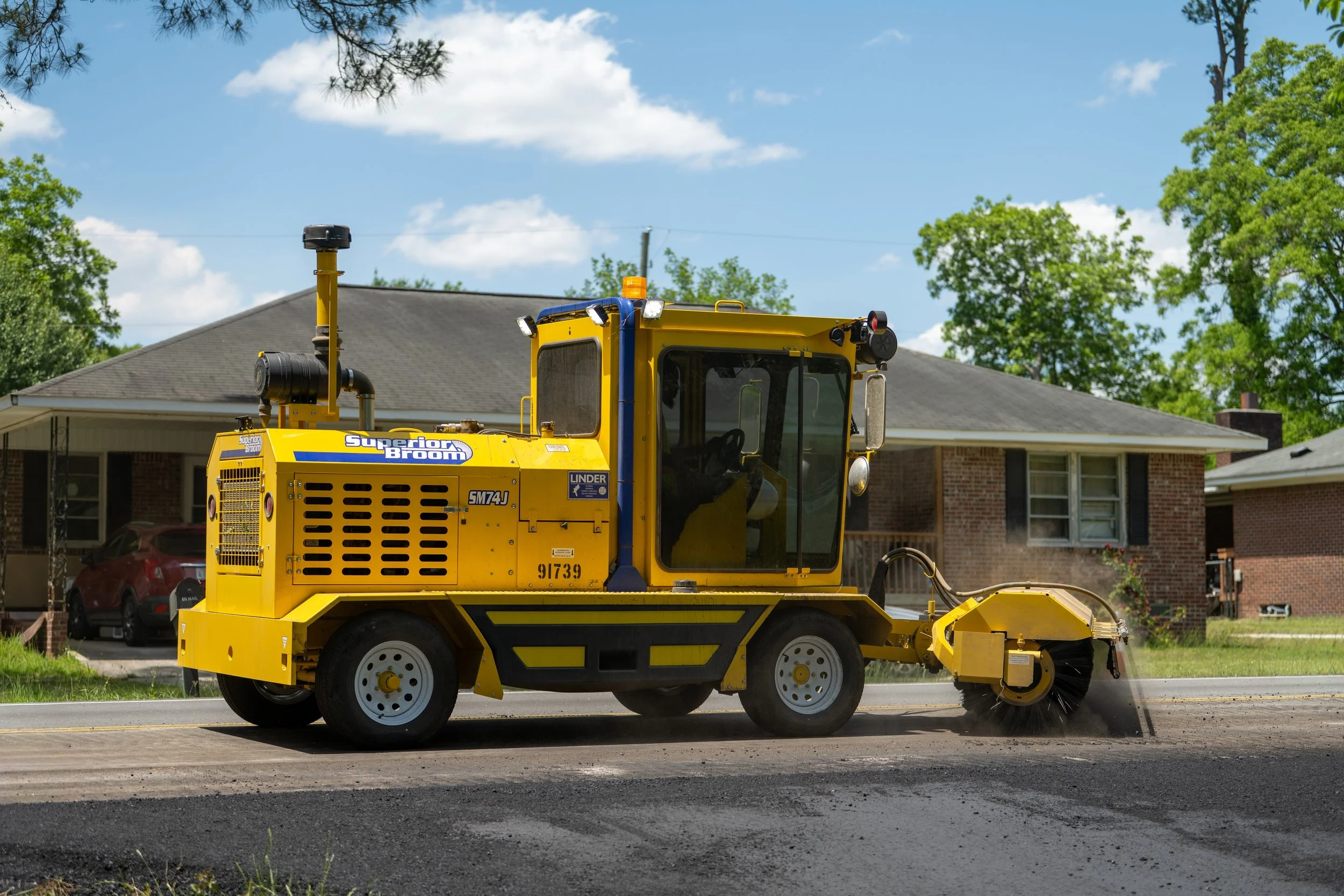 A yellow road sweeper vehicle cleaning a street in a residential neighborhood with houses and trees under a blue sky.