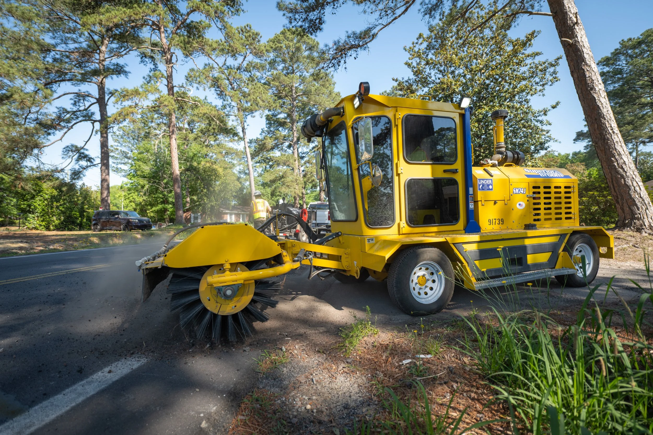 A yellow street sweeper cleaning a road surrounded by trees and greenery.