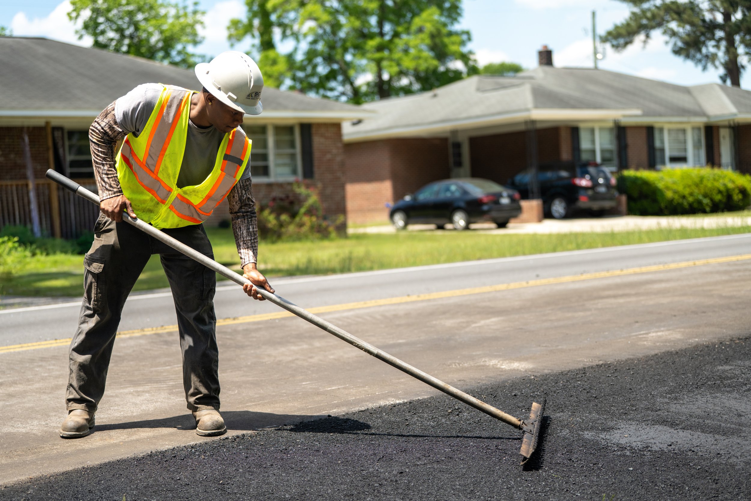 A construction worker wearing a white hard hat, high-visibility safety vest, and work boots, using a rake to level fresh asphalt on a street in a residential neighborhood.