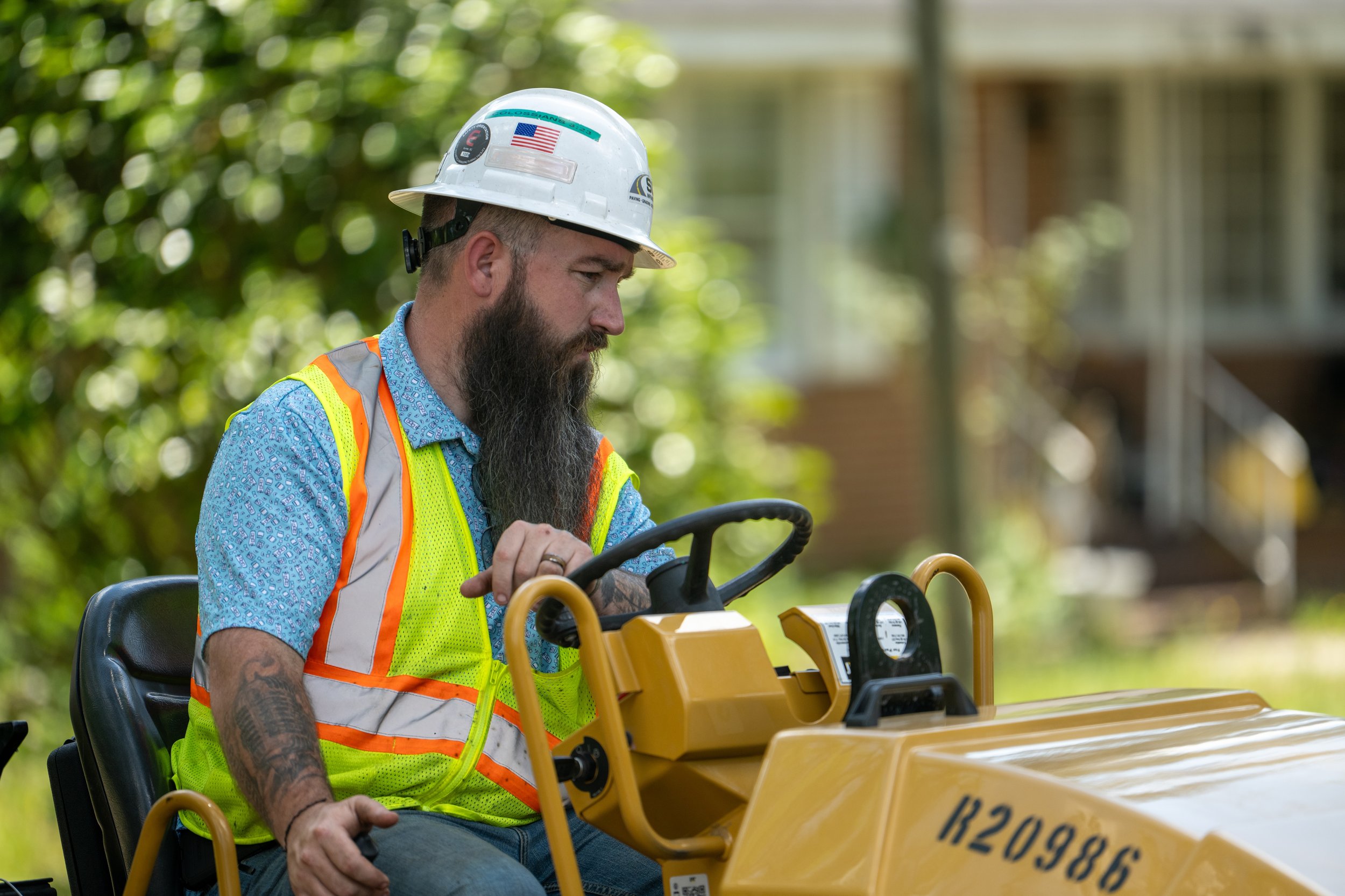 A man with a beard wearing a safety helmet with an American flag sticker and orange safety vest operating a yellow piece of machinery outdoors.