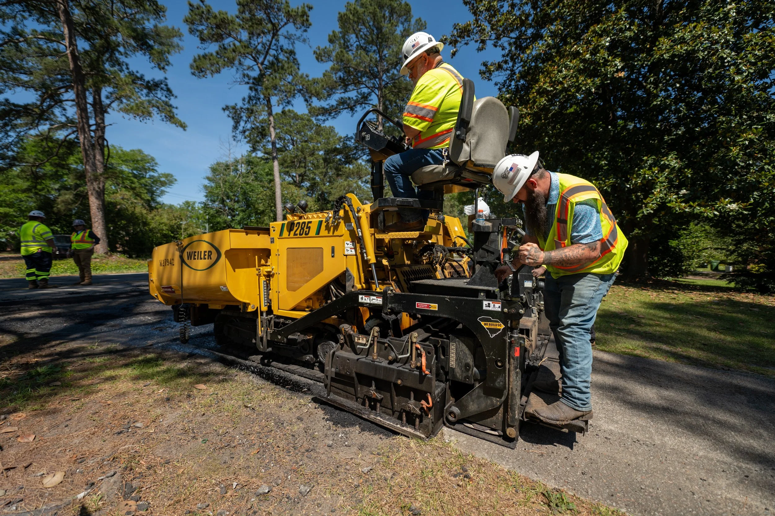 Workers in high-visibility vests and hard hats operating a road paving machine on a sunny day with trees and blue sky in the background.