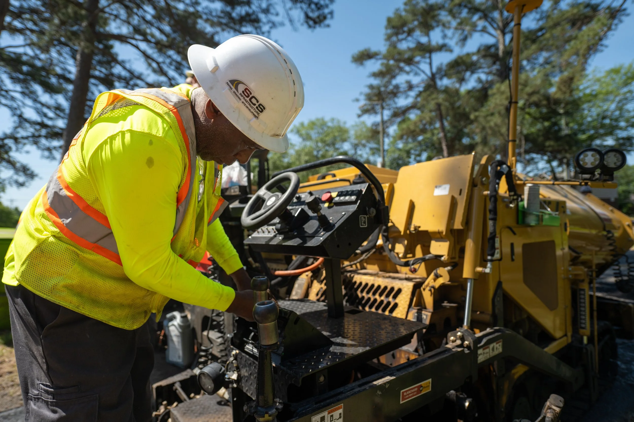 A construction worker wearing a white hard hat and yellow safety vest working on a yellow construction machine outdoors.
