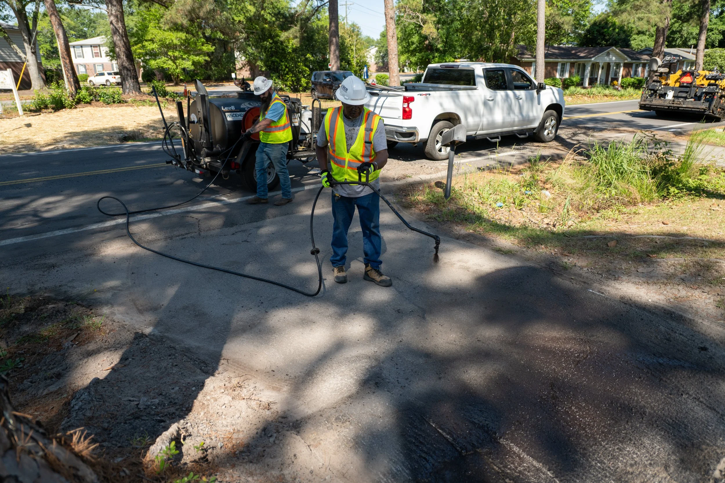 Two construction workers wearing safety vests and helmets working on road repair, with equipment, a white pickup truck, and a black utility vehicle nearby in a suburban neighborhood with trees and houses.