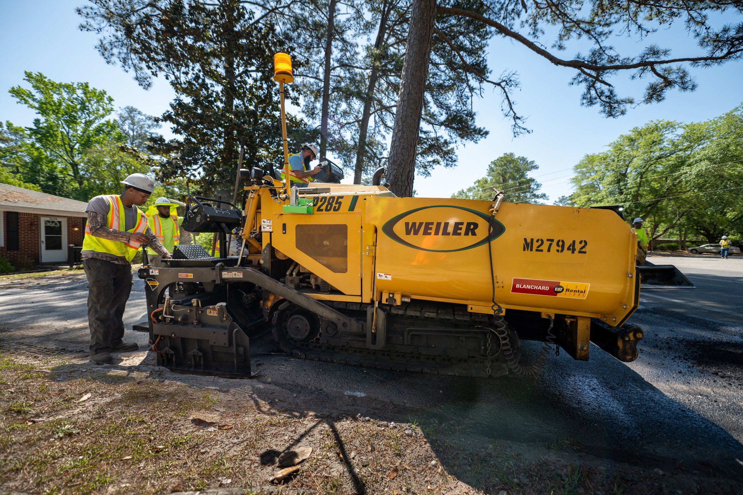 Construction workers operating a large yellow asphalt paving machine on a residential street, with trees and a house in the background.