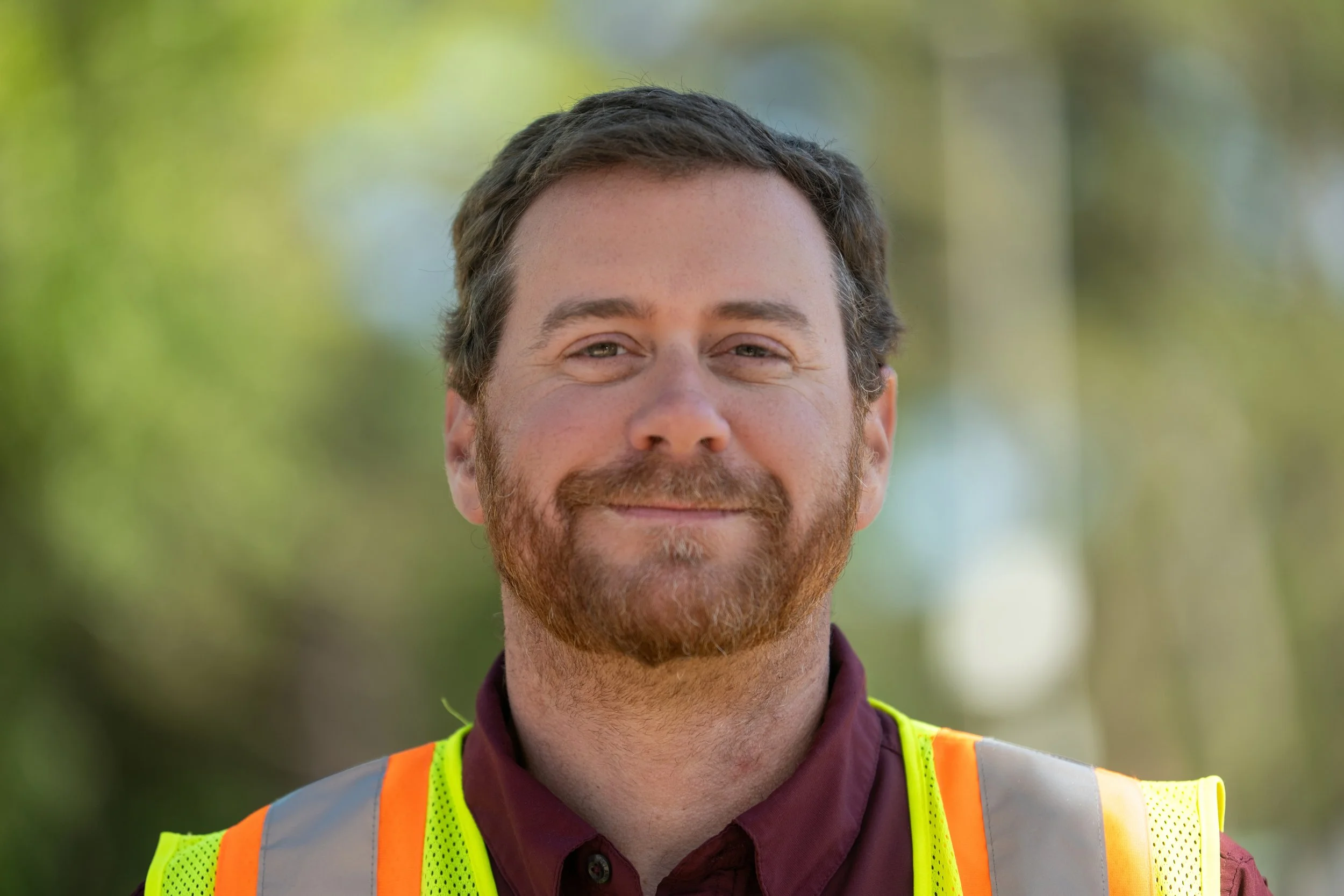 A man with a beard and short brown hair smiling outdoors, wearing a yellow safety vest over a maroon shirt, with a background of green trees.