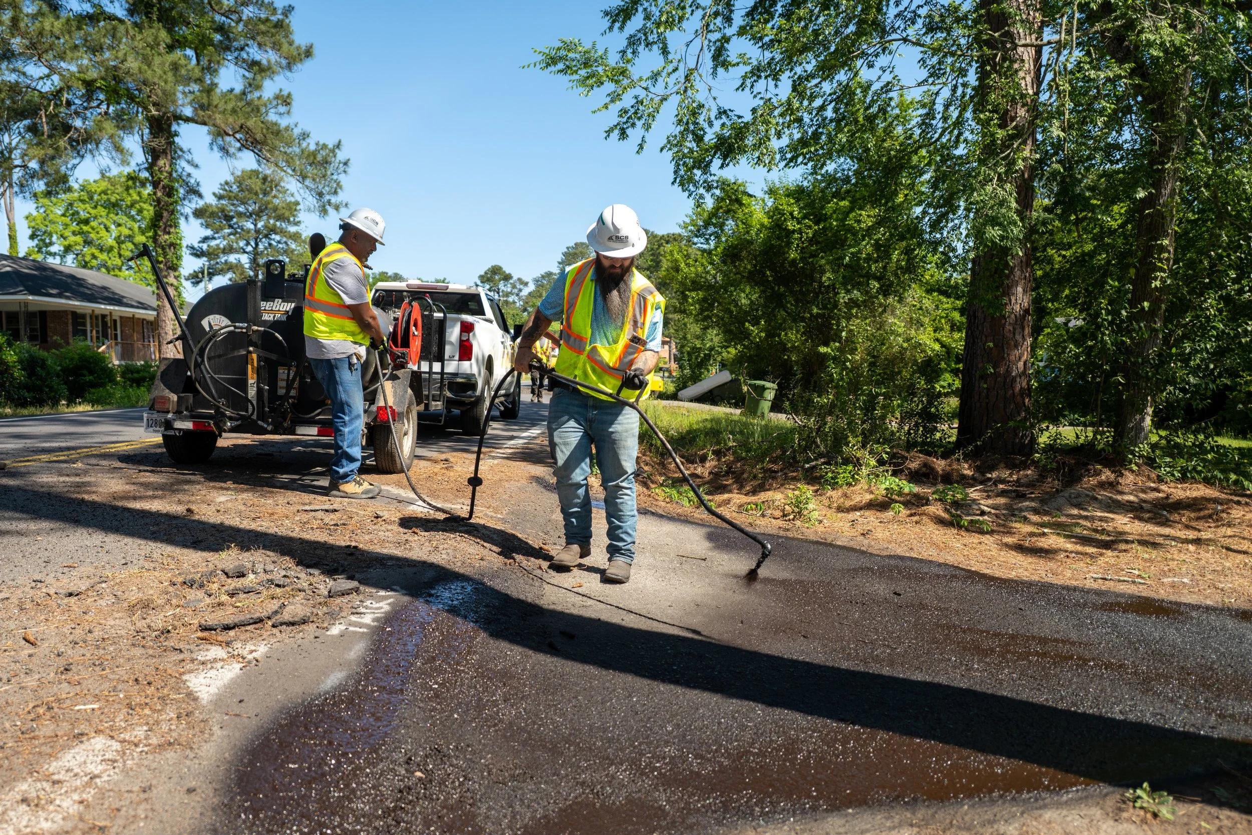 Two construction workers wearing safety vests and hard hats working on asphalt patching on a street, with trees and houses in the background.