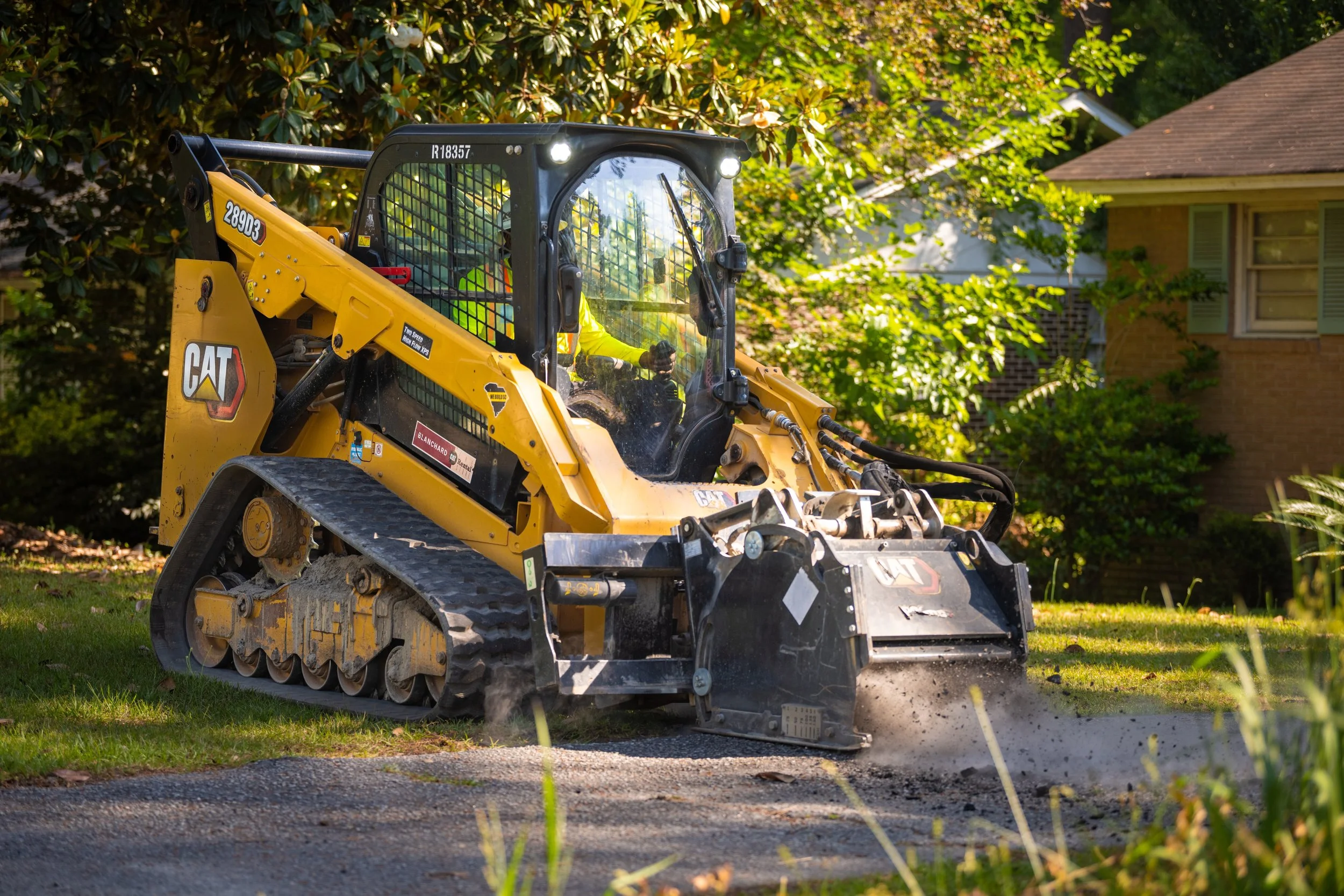 A yellow Caterpillar skid steer loader working on a residential yard, with trees and house in the background.
