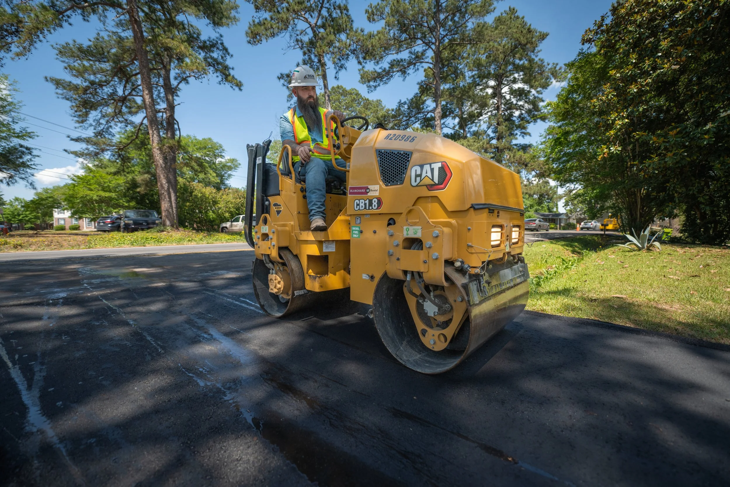 A construction worker operating a yellow CAT CB1.8 vibratory roller compactor on a paved road surrounded by trees and parked cars in a residential area.