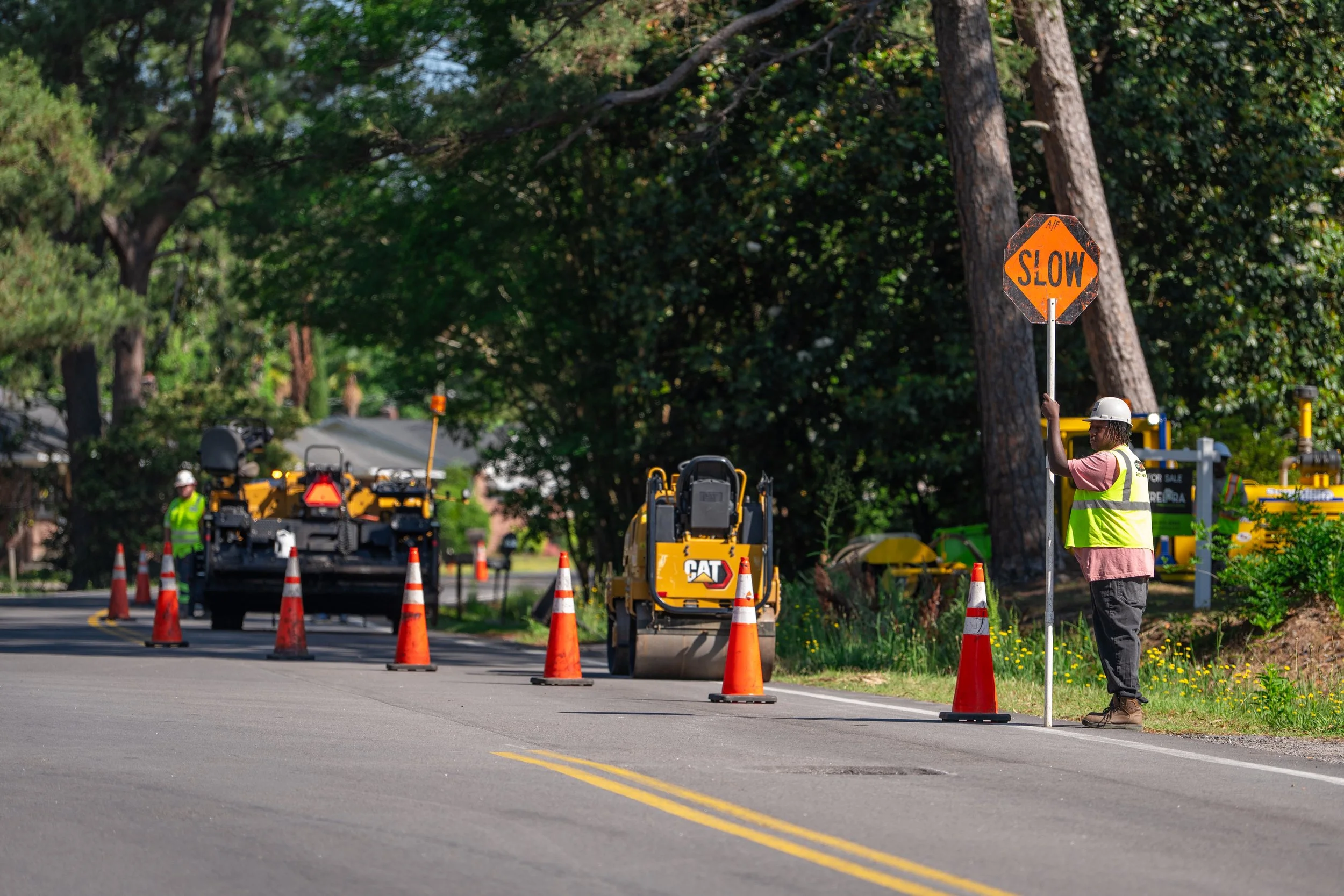 Construction workers and equipment on a residential street with a 'slow' sign held by a worker wearing a white safety helmet, high-visibility vest, pink shirt, and gray pants. Orange traffic cones mark the roadwork area, surrounded by trees and greenery.