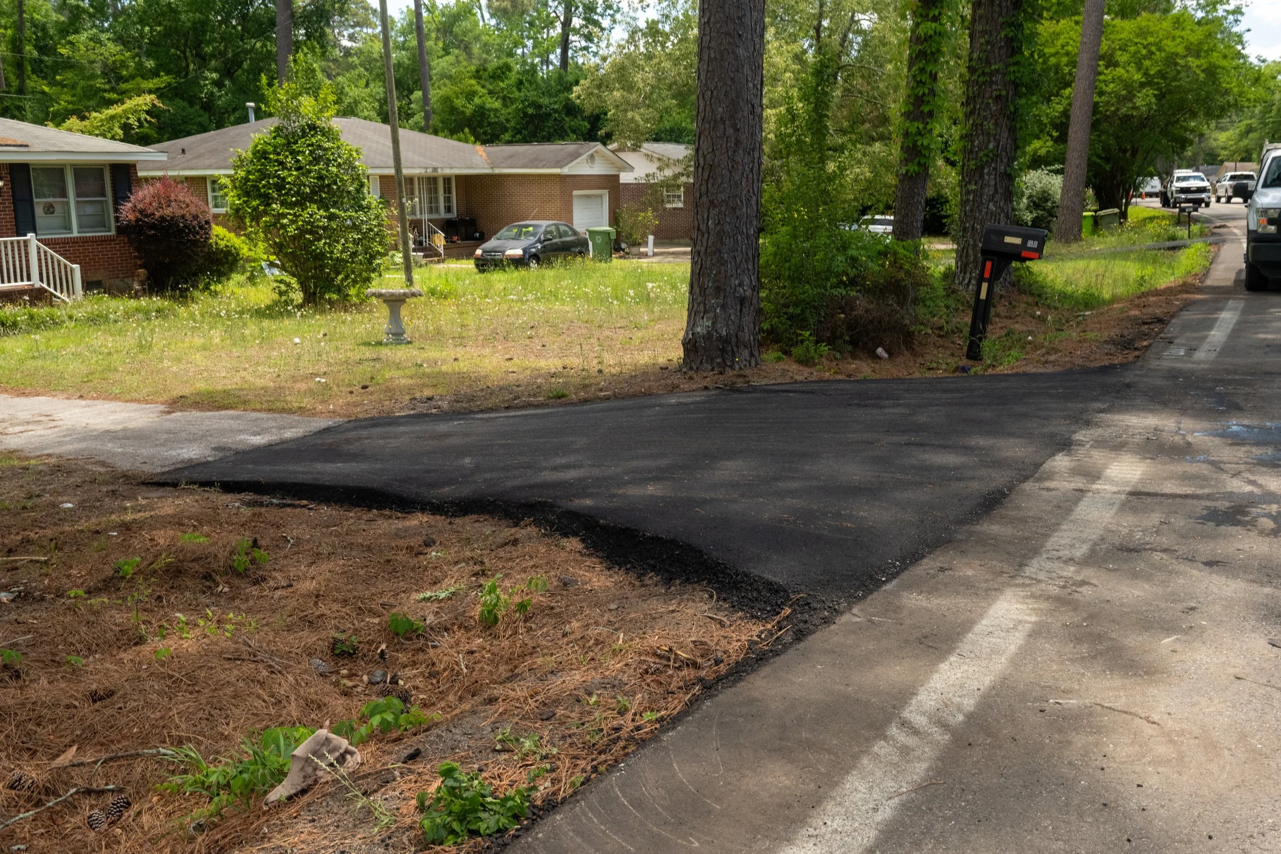 A residential neighborhood with a freshly paved driveway, trees, grass, and suburban houses in the background.