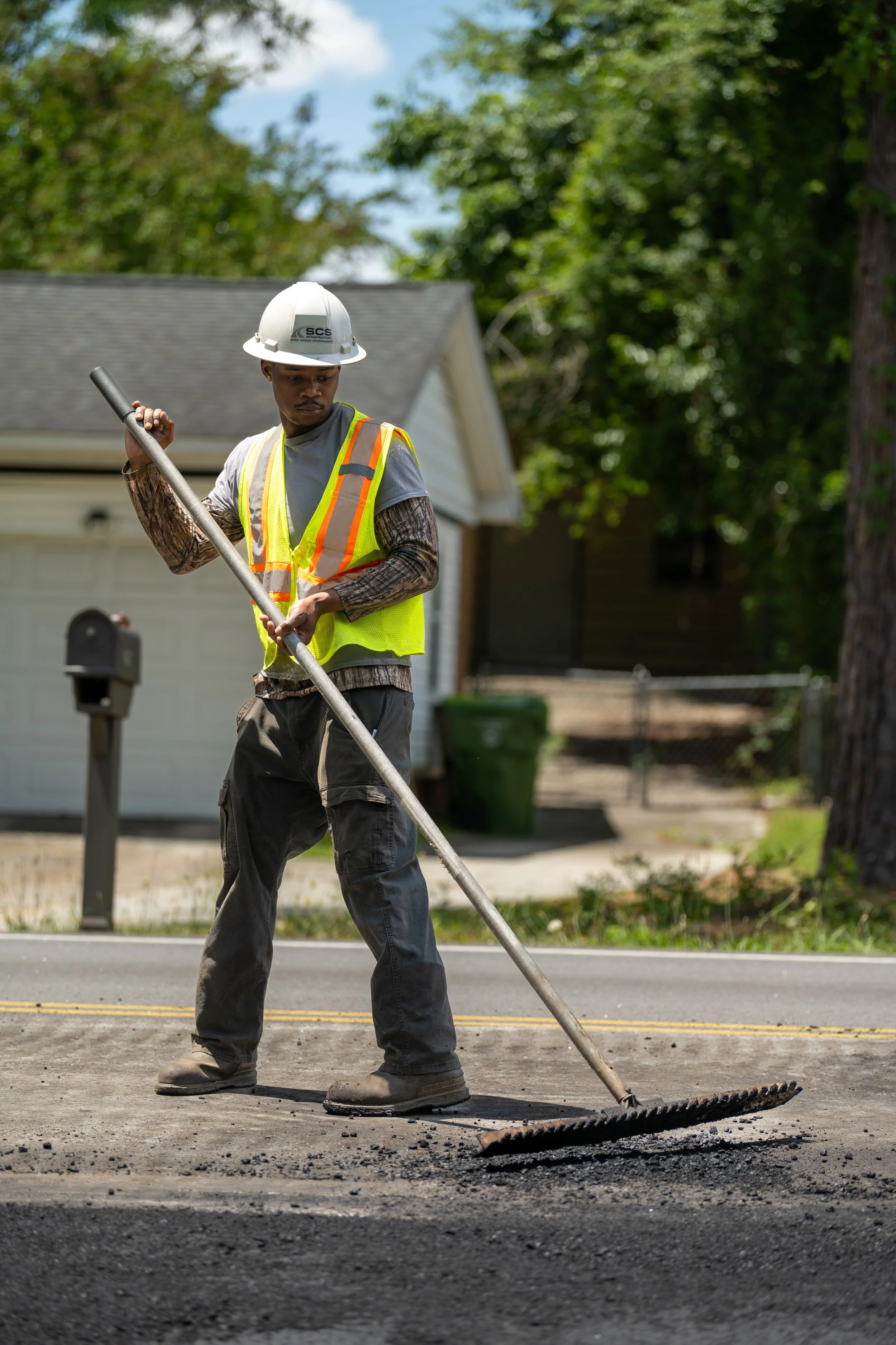A worker wearing a safety helmet and reflective vest is using a rake to level blacktop on a street.