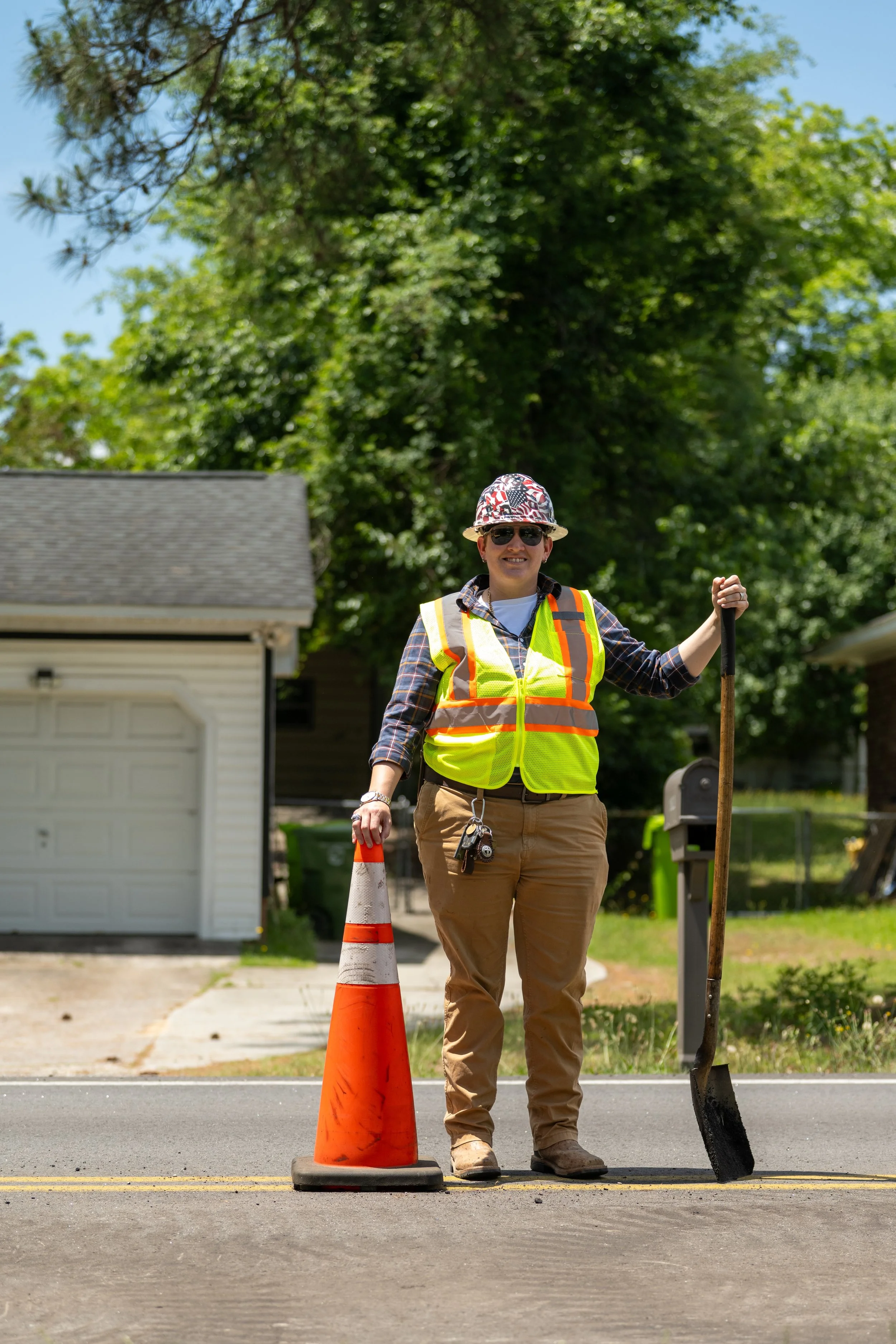 A person wearing a safety vest, hat, and sunglasses stands on a road holding a shovel, with orange traffic cones nearby, and a residential building in the background under a clear blue sky.