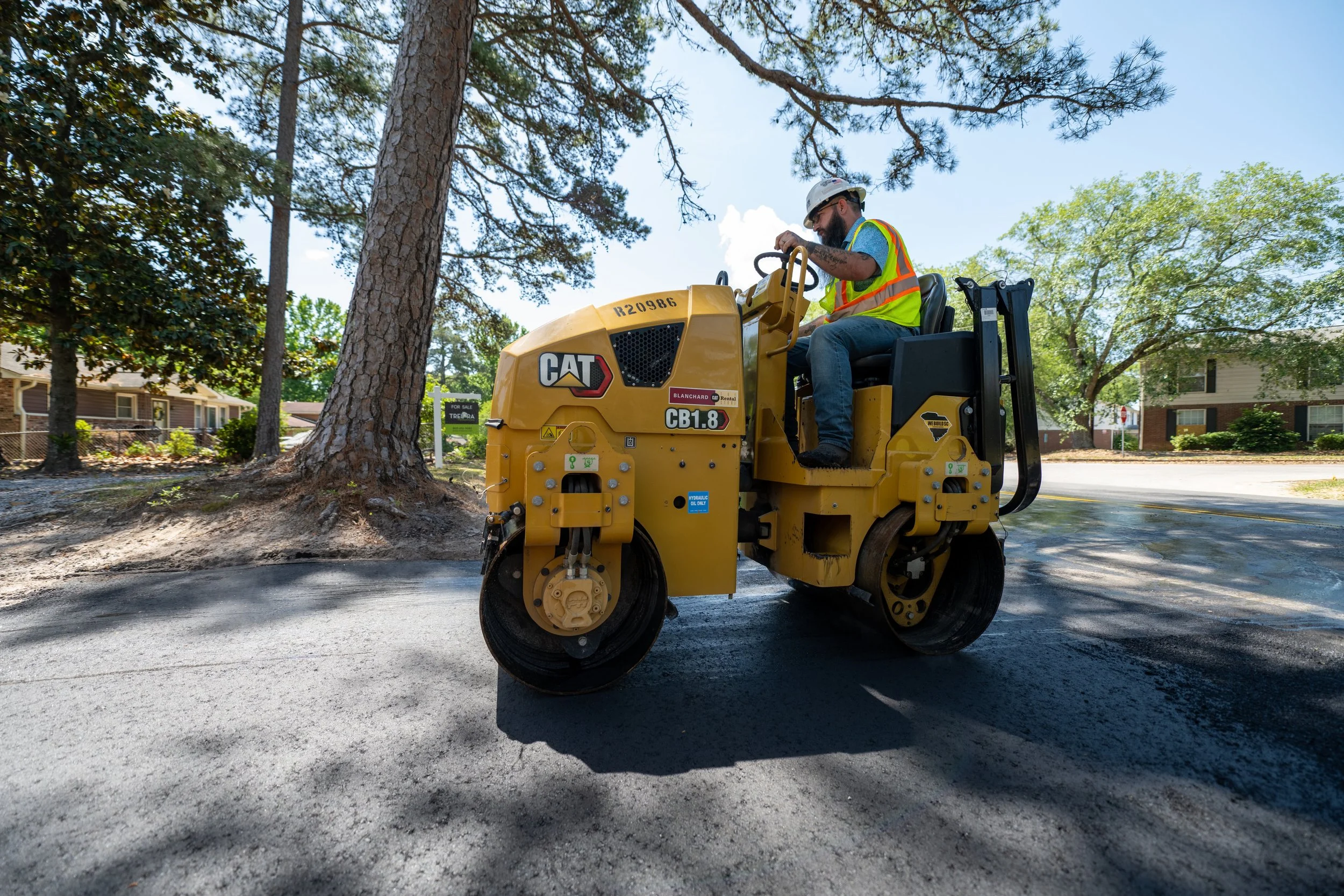 A construction worker operating a yellow CAT CB1.8 compact roller on a freshly paved road in a residential neighborhood with trees and houses in the background.