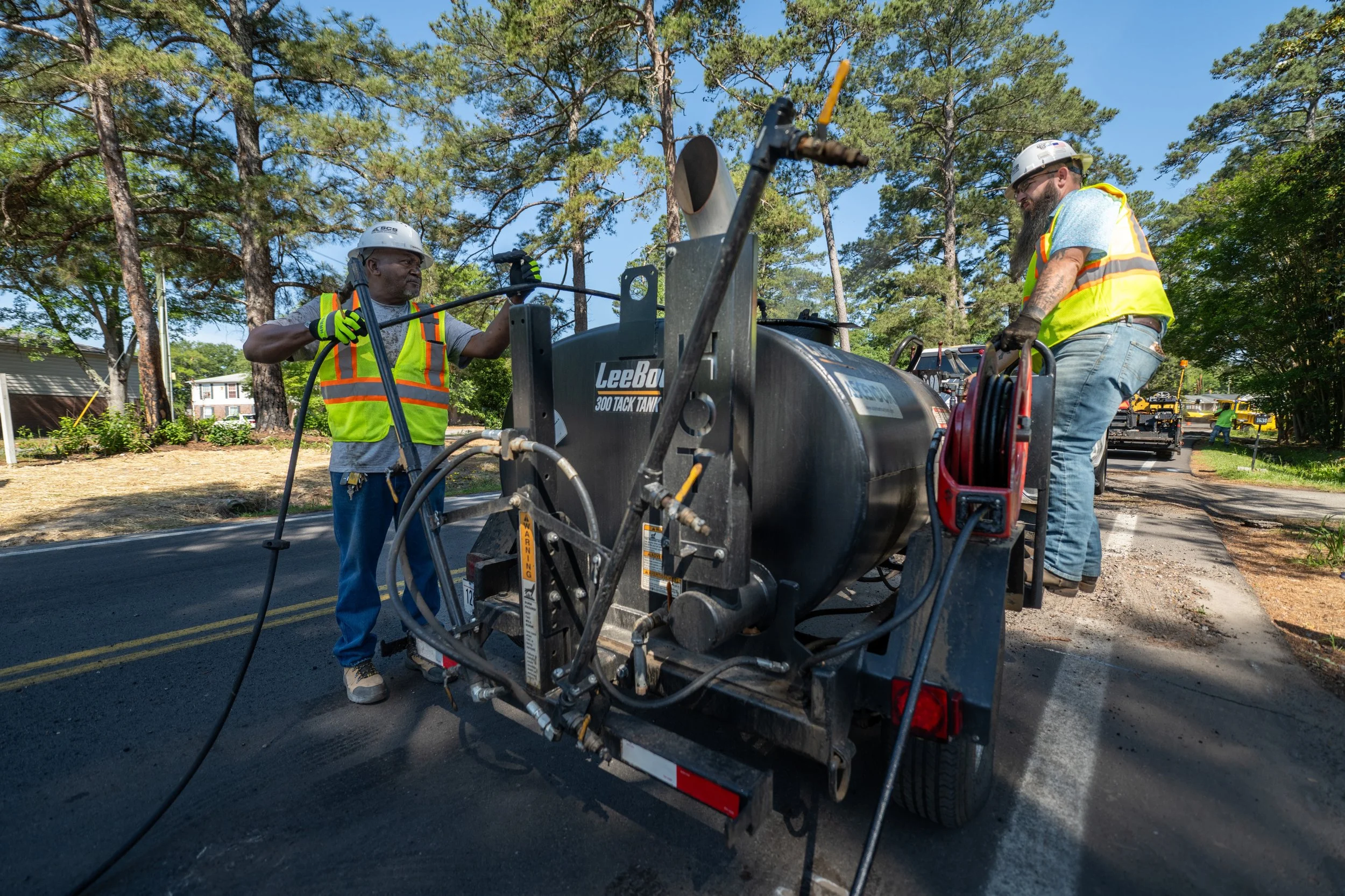 Two construction workers wearing yellow safety vests and helmets operating a large road paving machine on a street surrounded by trees and residential houses.