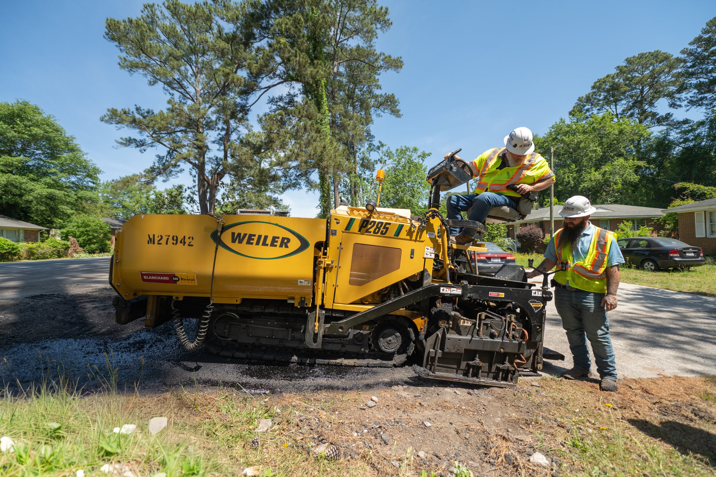 Two workers in safety vests and helmets operate a yellow road paving machine on a residential street, with trees, houses, and parked cars in the background, under a clear blue sky.
