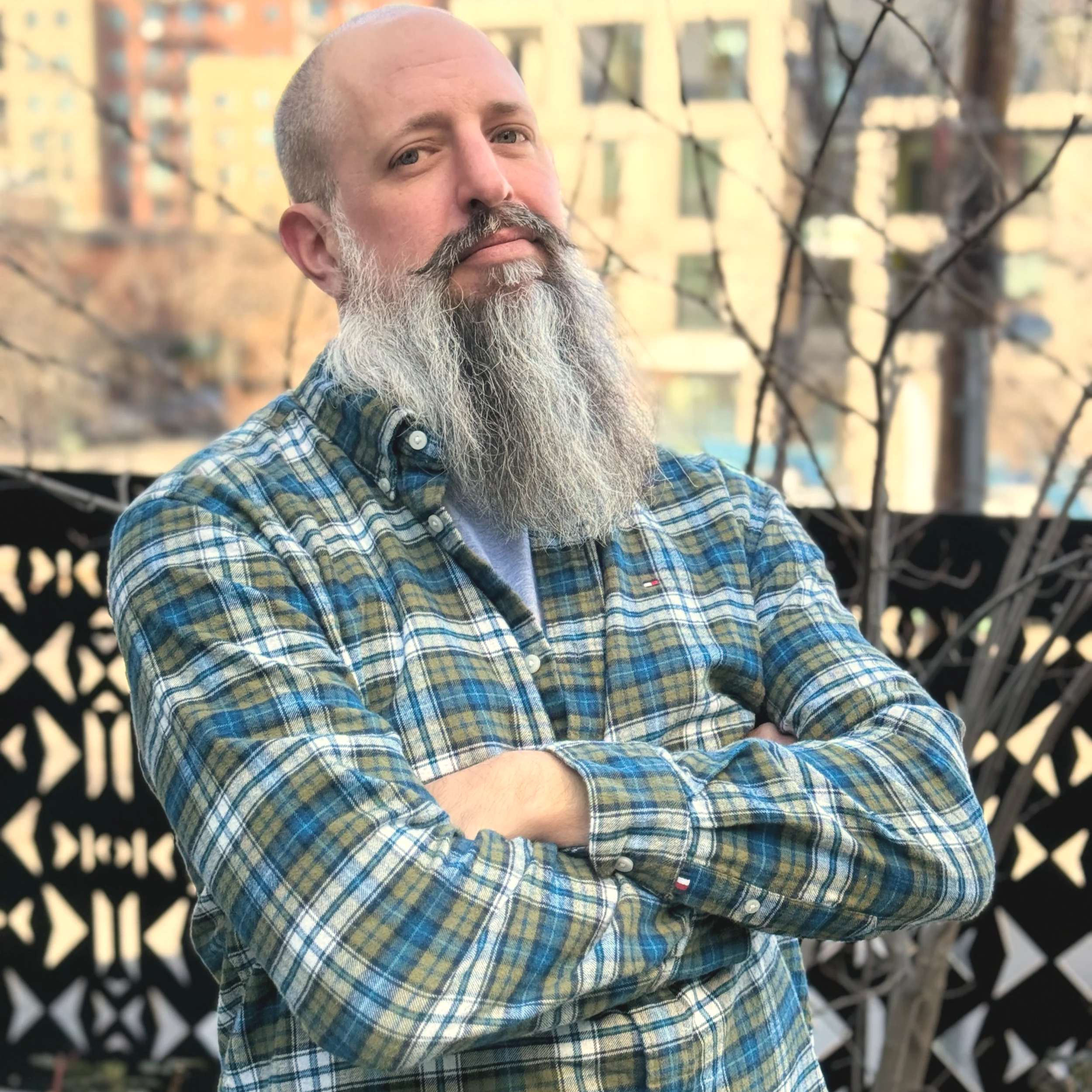 A middle-aged man with a gray beard and bald head wearing a plaid shirt, standing outdoors with buildings and leafless trees in the background.