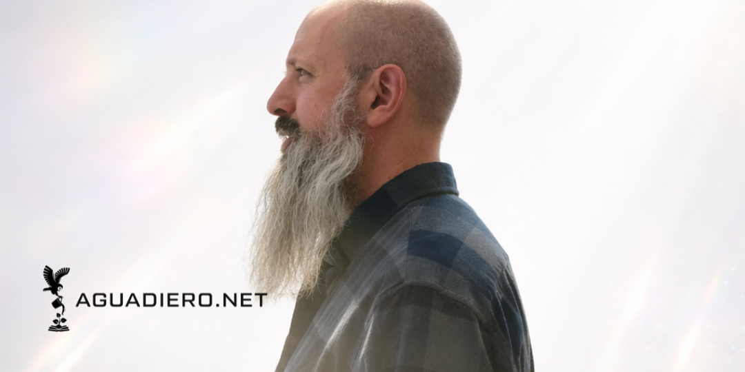 Side profile of a man with a beard and shaved head wearing a dark plaid shirt against a light, cloudy background with rainbow-like light effects.