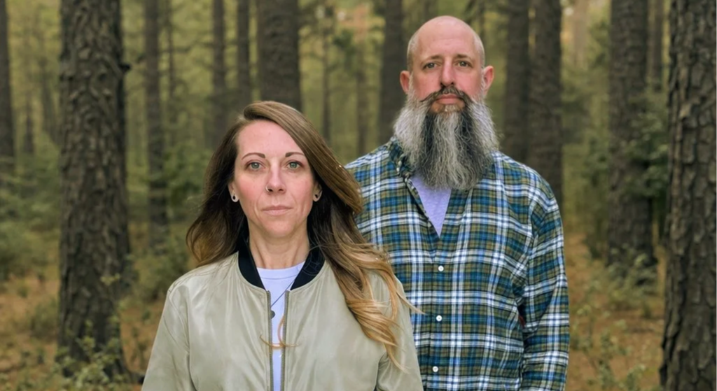 A woman and a man stand outdoors in a forest with tall trees, looking serious at the camera.