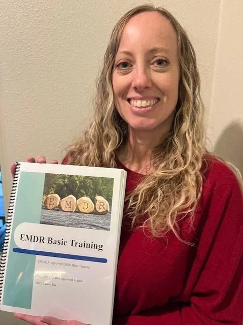 A woman with long blonde curly hair wearing a red sweater holding a course book titled 'EMDR Basic Training'.