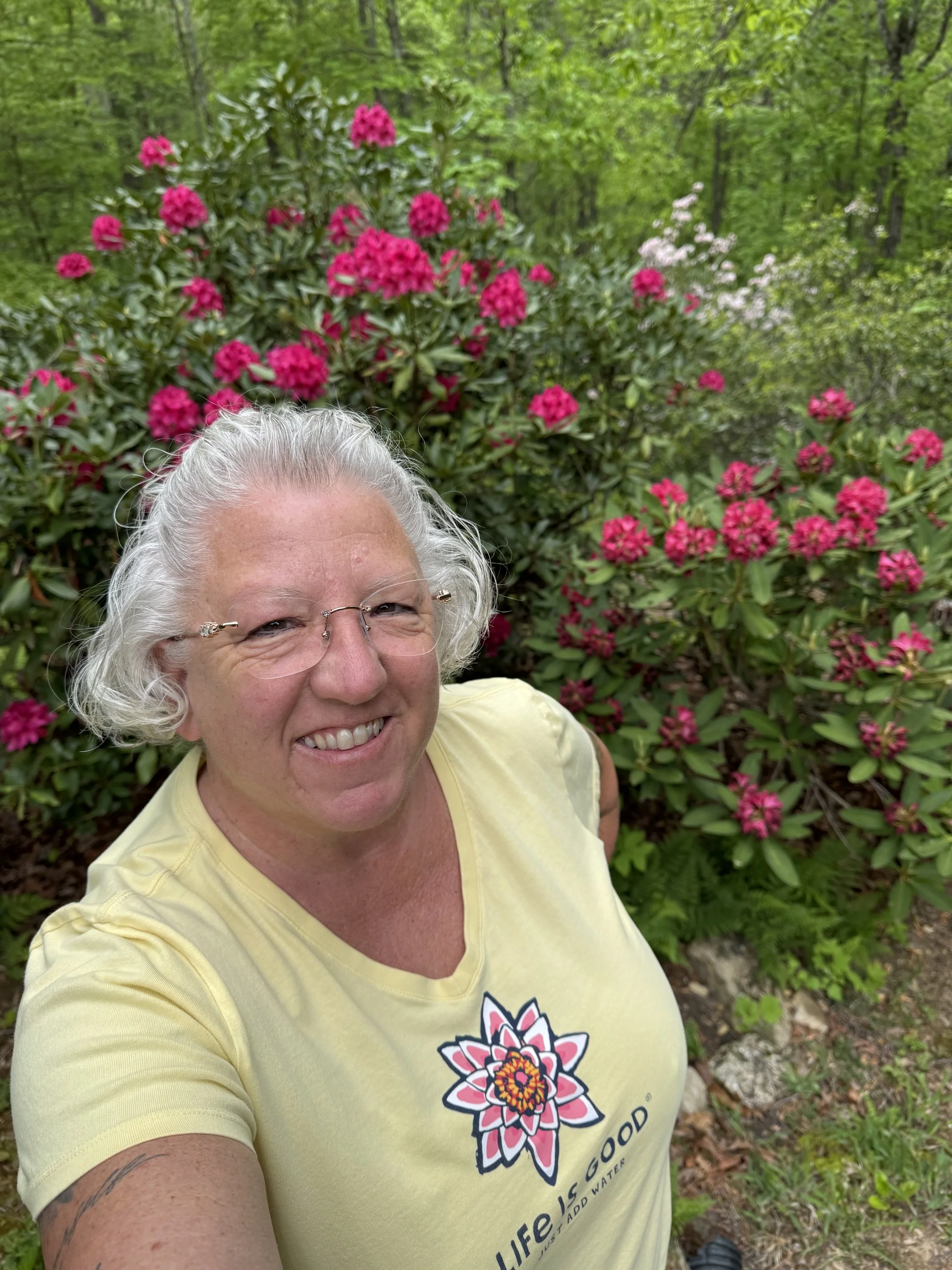 A smiling woman with gray hair and glasses taking a selfie outdoors in front of a bush with pink flowers and green leaves.