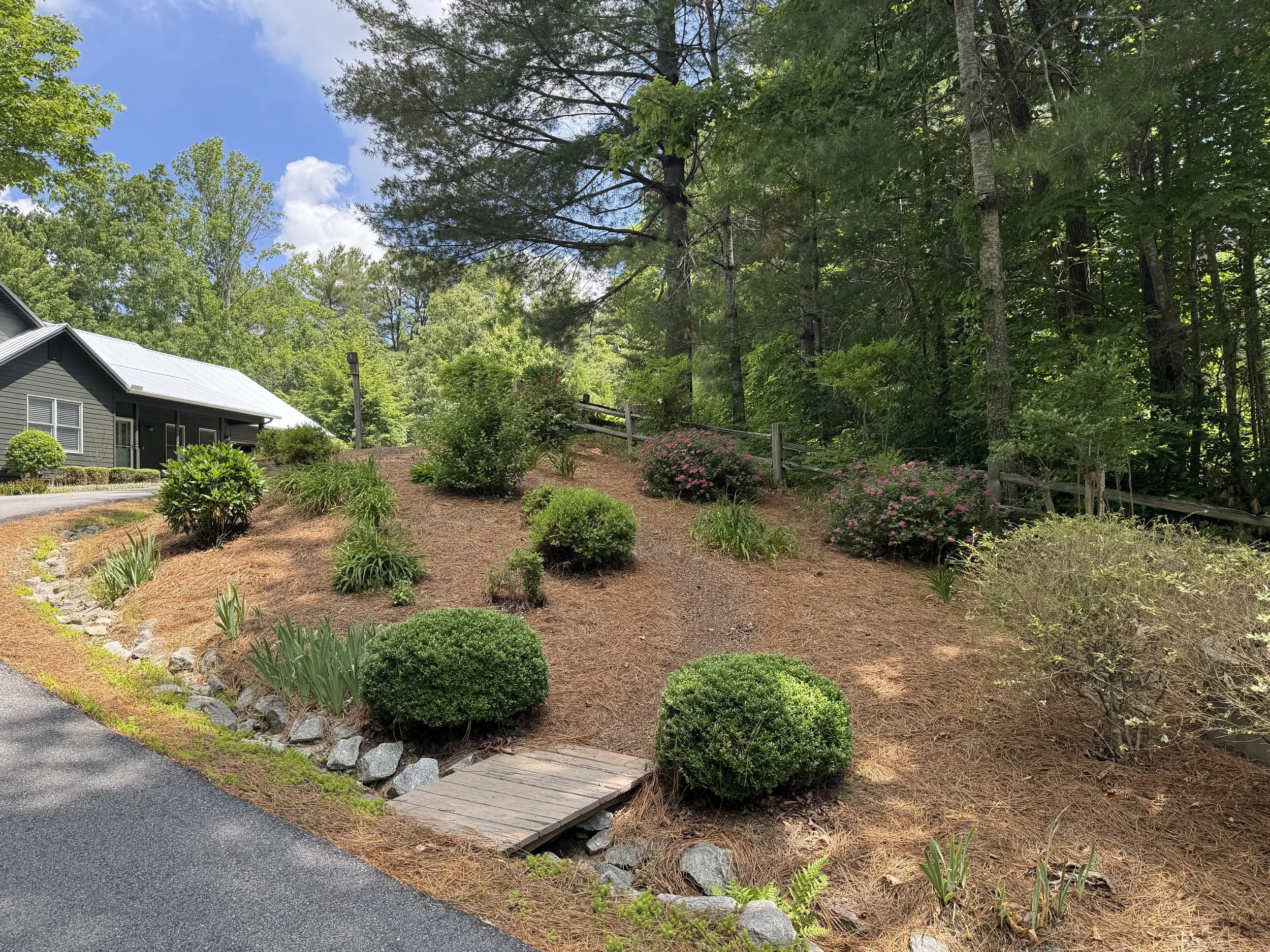 A garden with a paved walkway, wooden bridge, and landscaped bushes and flowers amidst tall green trees under a partly cloudy blue sky.