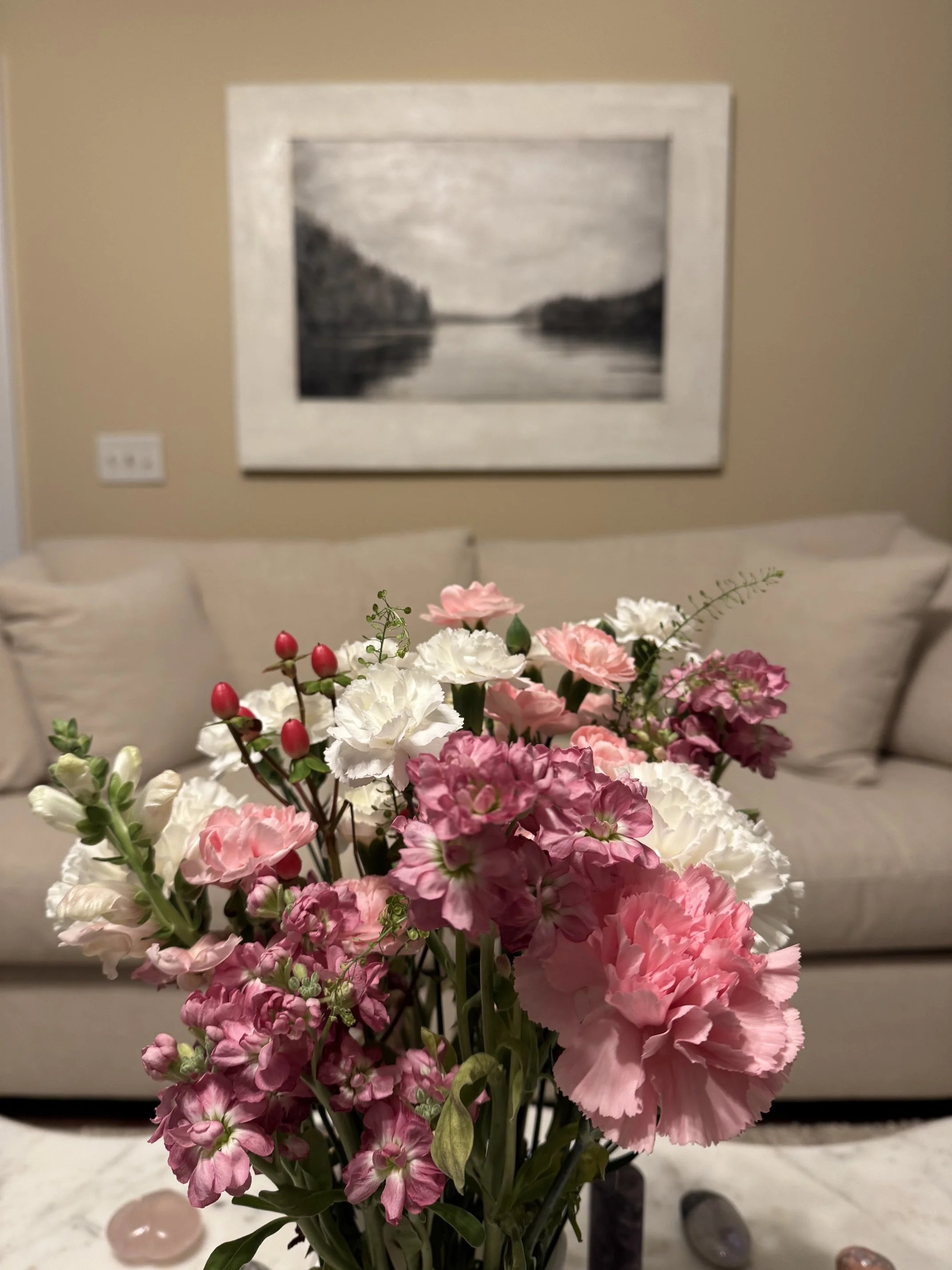 A bouquet of pink and white flowers on a table in the foreground, with a beige wall, a black and white landscape photograph, a white sofa, and decorative stones in a pink container in the background.