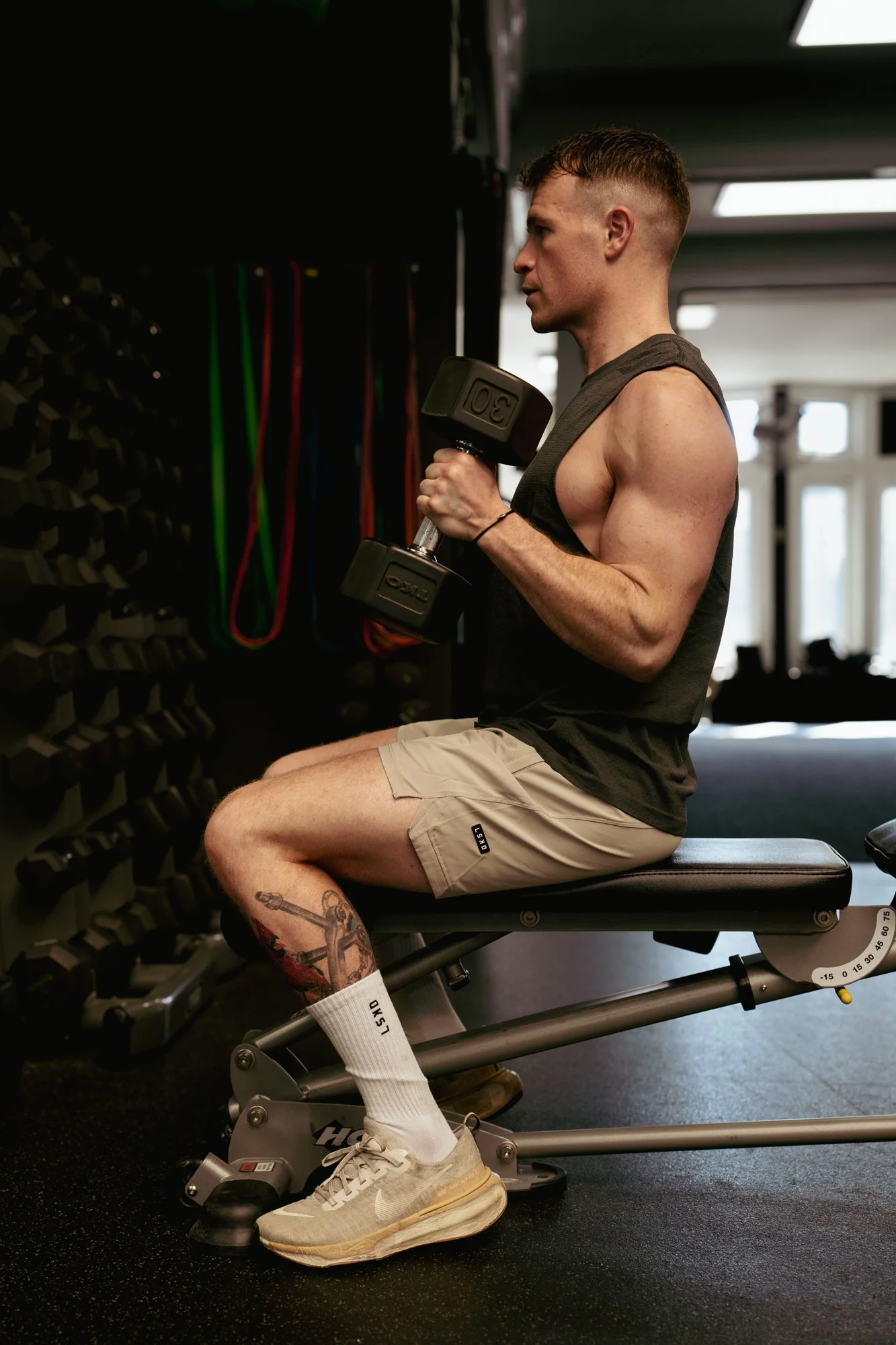 A man sitting on a workout bench in a gym, holding a dumbbell in one hand, with resistance bands hanging on the wall nearby.