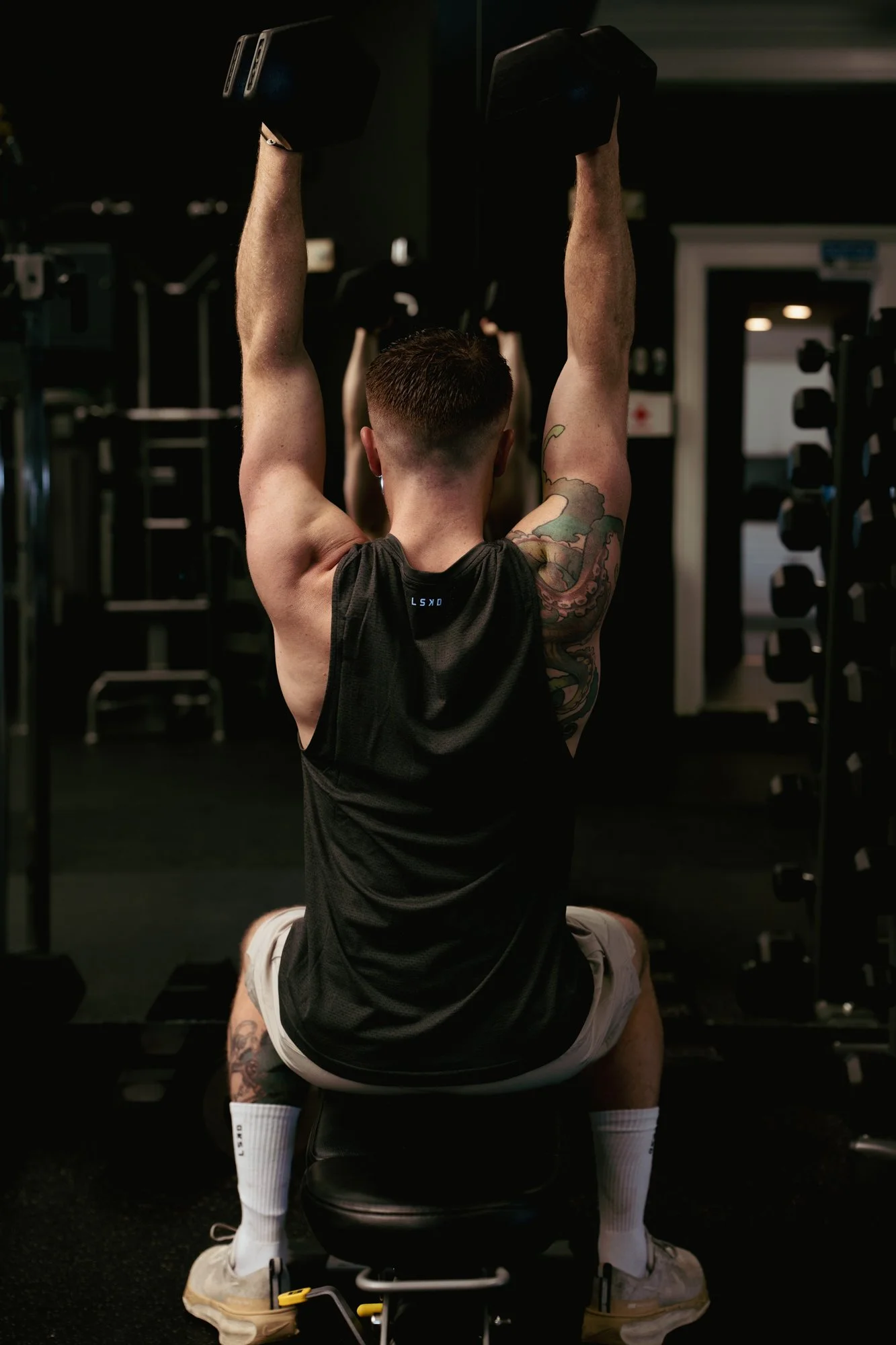 A man doing a shoulder press exercise with dumbbells in a gym, seated on a workout bench, viewed from behind.