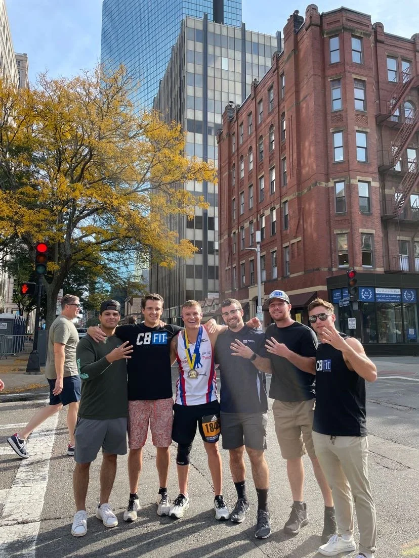 Group of six young men standing on city street corner, smiling, some with medals, in front of tall buildings and a leafless tree, during daytime.