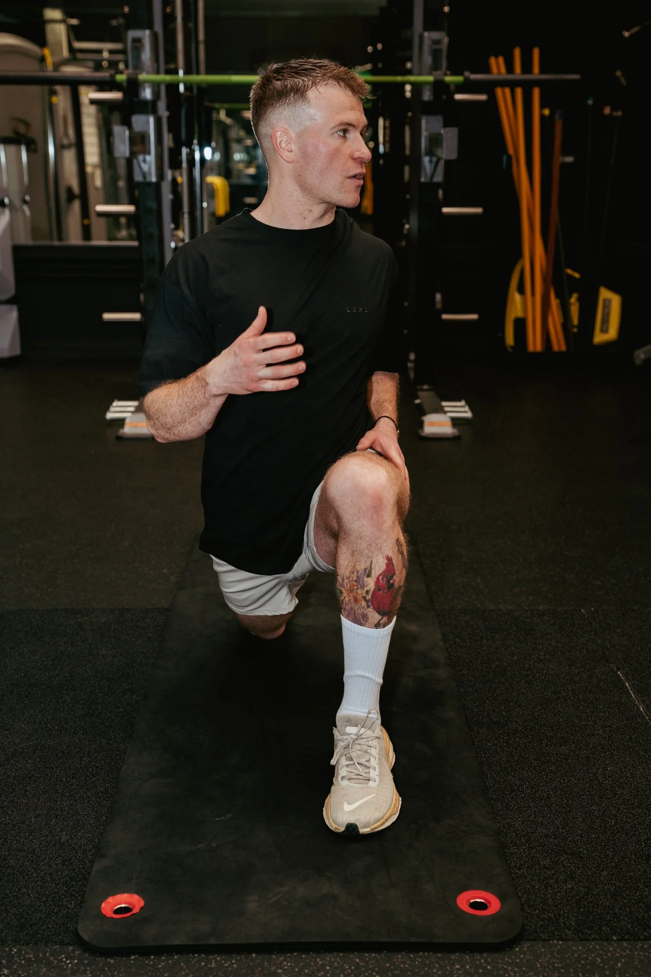 A man is doing a lunge exercise on a black workout mat in a gym, wearing a black shirt, beige shorts, white socks, and beige sneakers. The gym has black walls, workout equipment, and yellow resistance bands in the background.