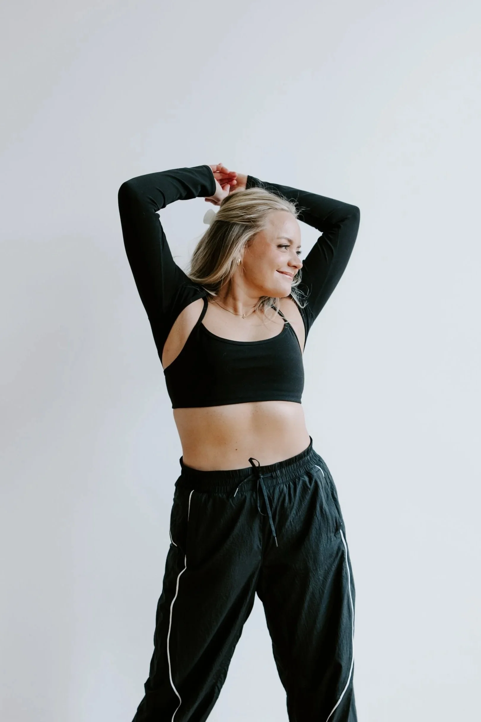 A woman in a black sports bra and black track pants smiling with her arms raised behind her head, standing against a plain white background.