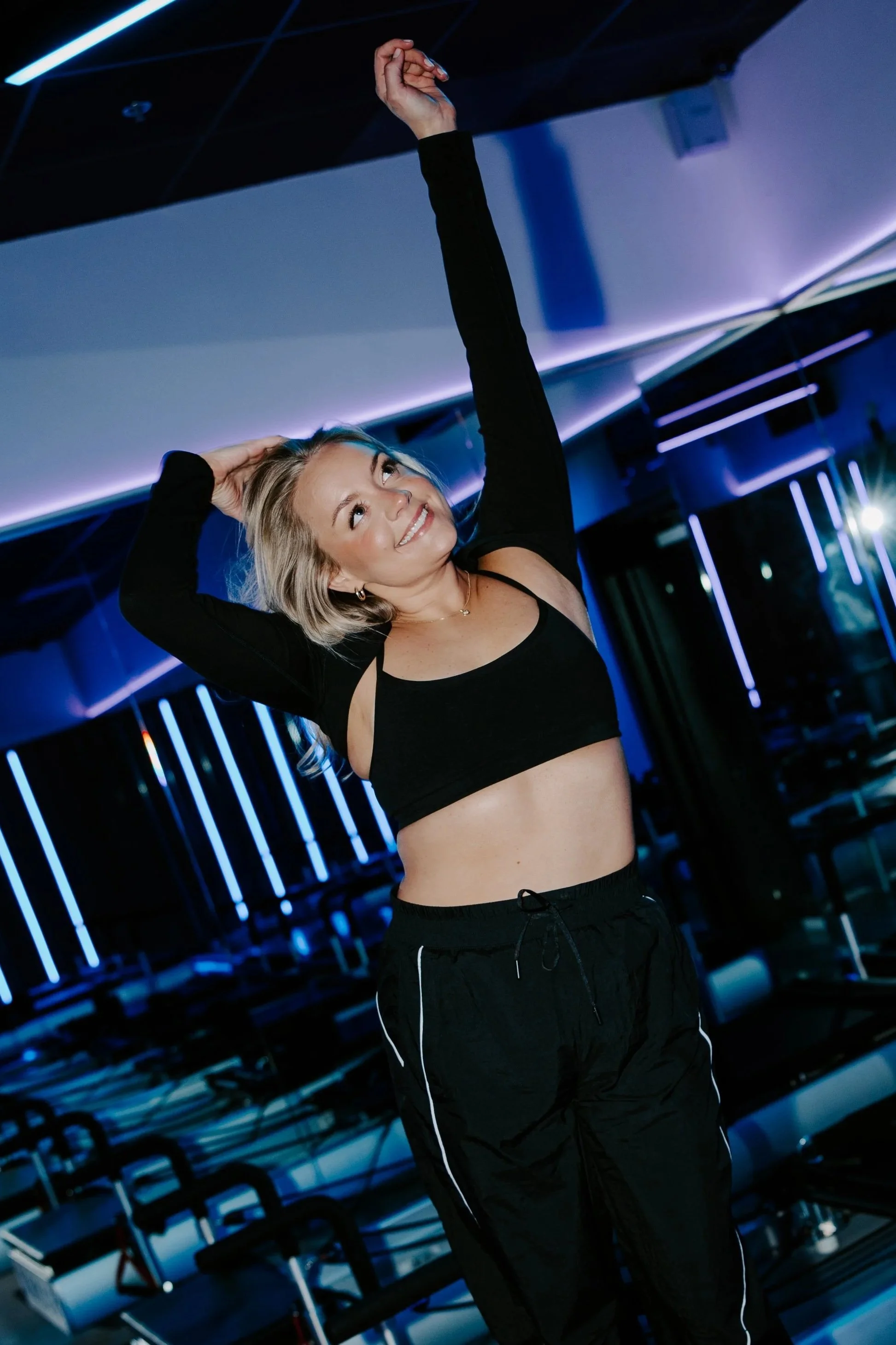 Young woman with blonde hair exercising in a gym with blue and purple lighting, wearing a black sports bra and black track pants.