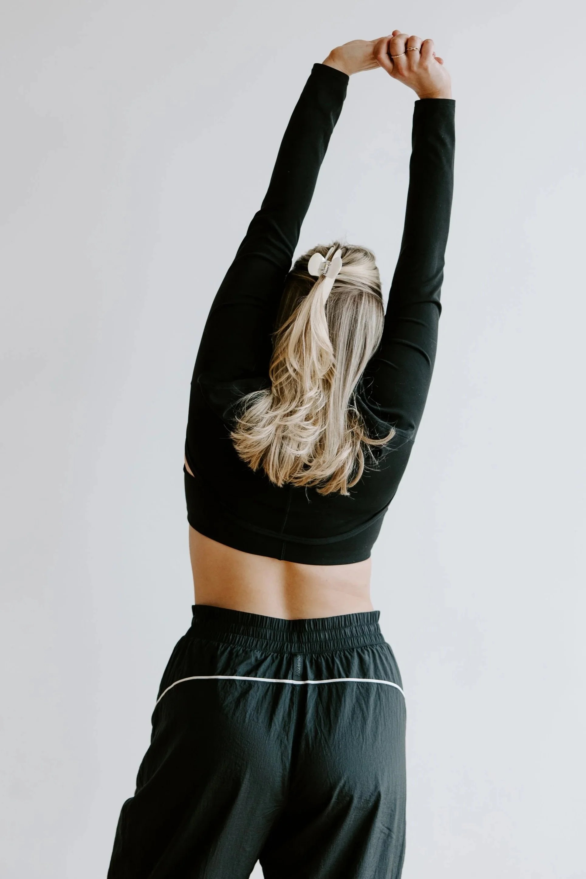 Person wearing black athletic shorts and a black long sleeve shirt, stretching arms overhead, and holding their hair with hair clips, against a plain white background.