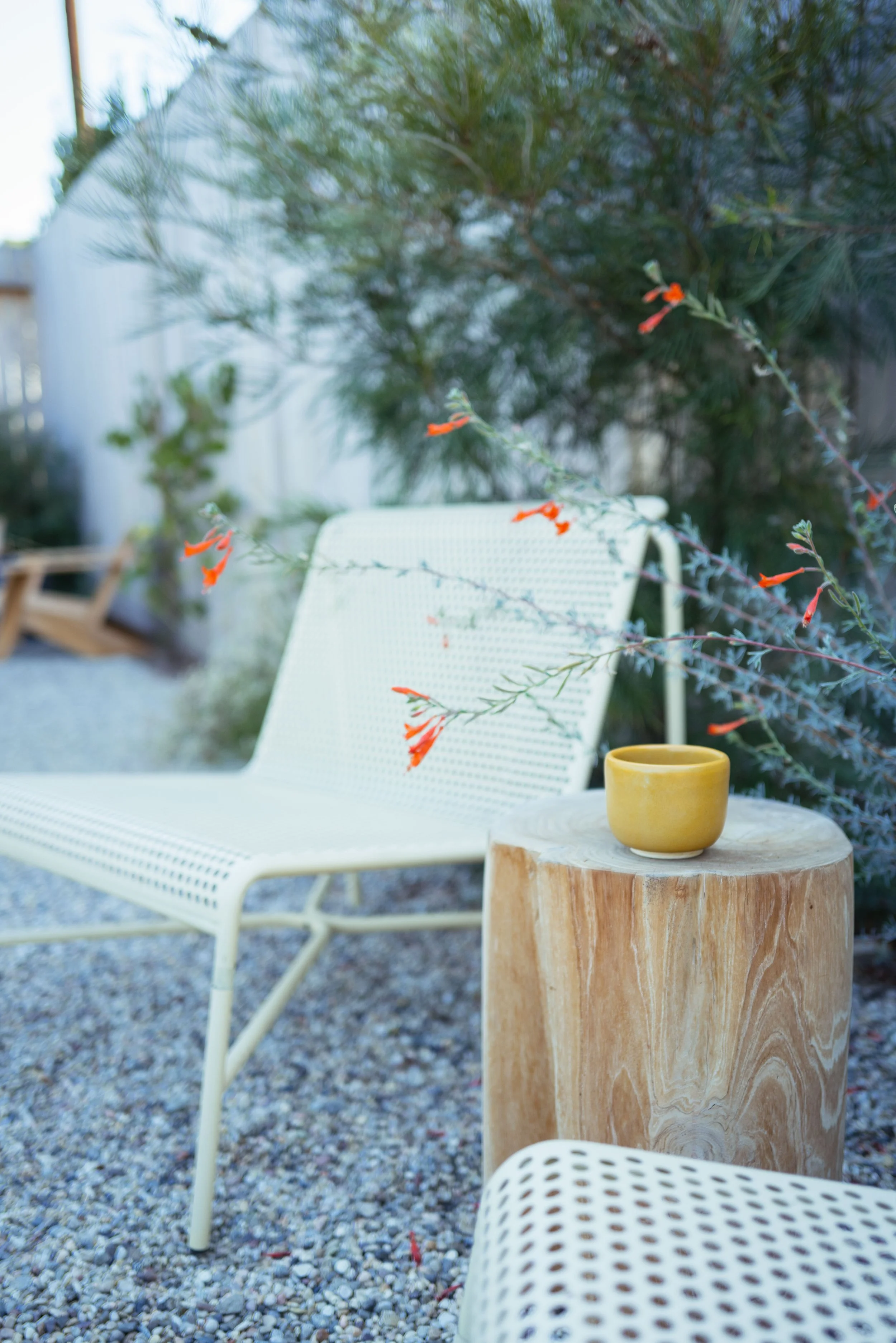 An outdoor seating area with a white mesh chair, a small round wood table with a yellow ceramic cup, a gravel ground, and green plants in the background.