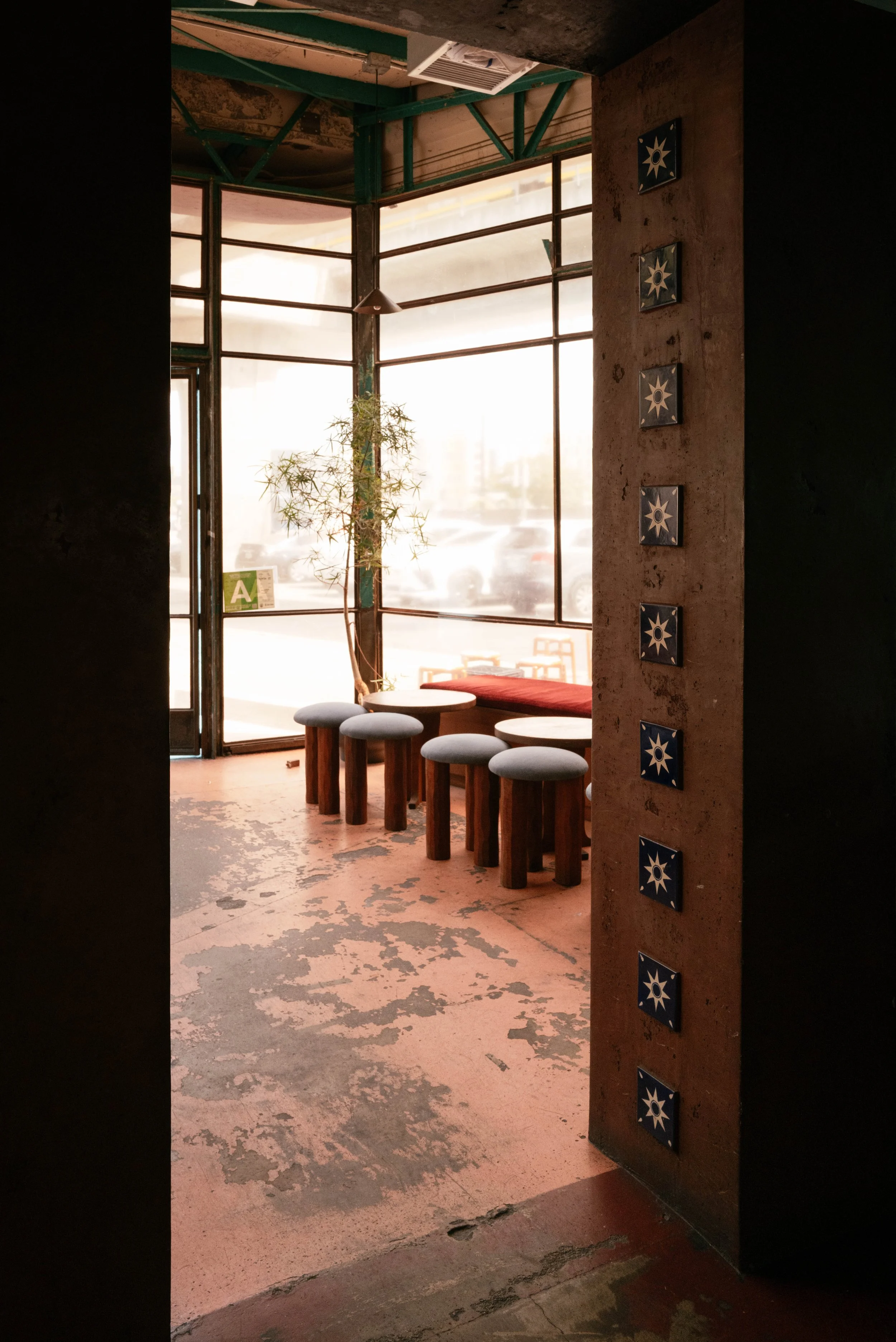 Interior view of a rustic cafe or restaurant with large windows, a small round table, five padded stools, a potted plant, and decorative tiles on a wall. The floor and some walls are worn and peeling.