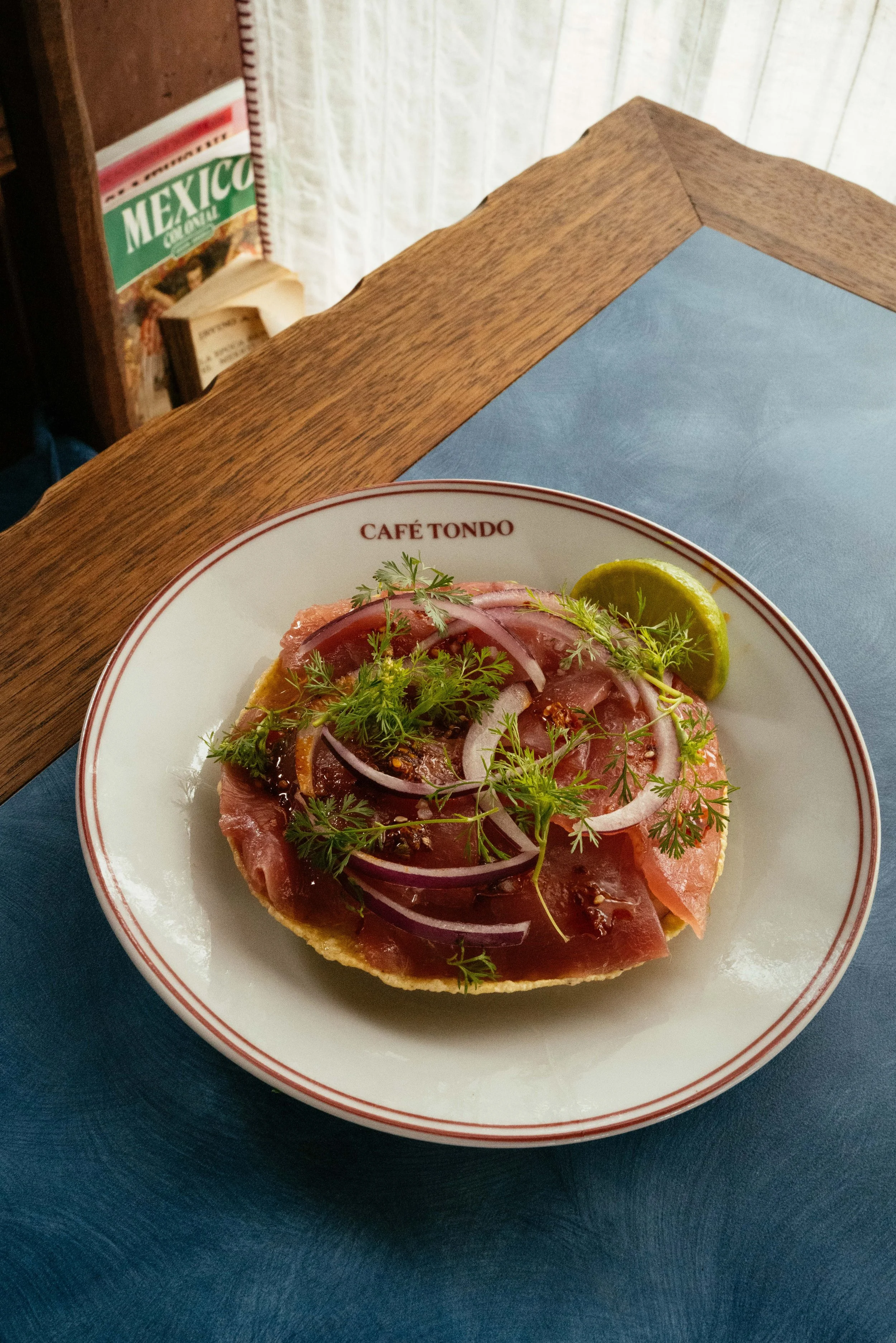 A plate of carne asada tostada topped with sliced onions, cilantro, and a lime wedge. The plate has a red rim and the words 'Café Tondo' on it, placed on a dark blue placemat with a wooden table in the background.