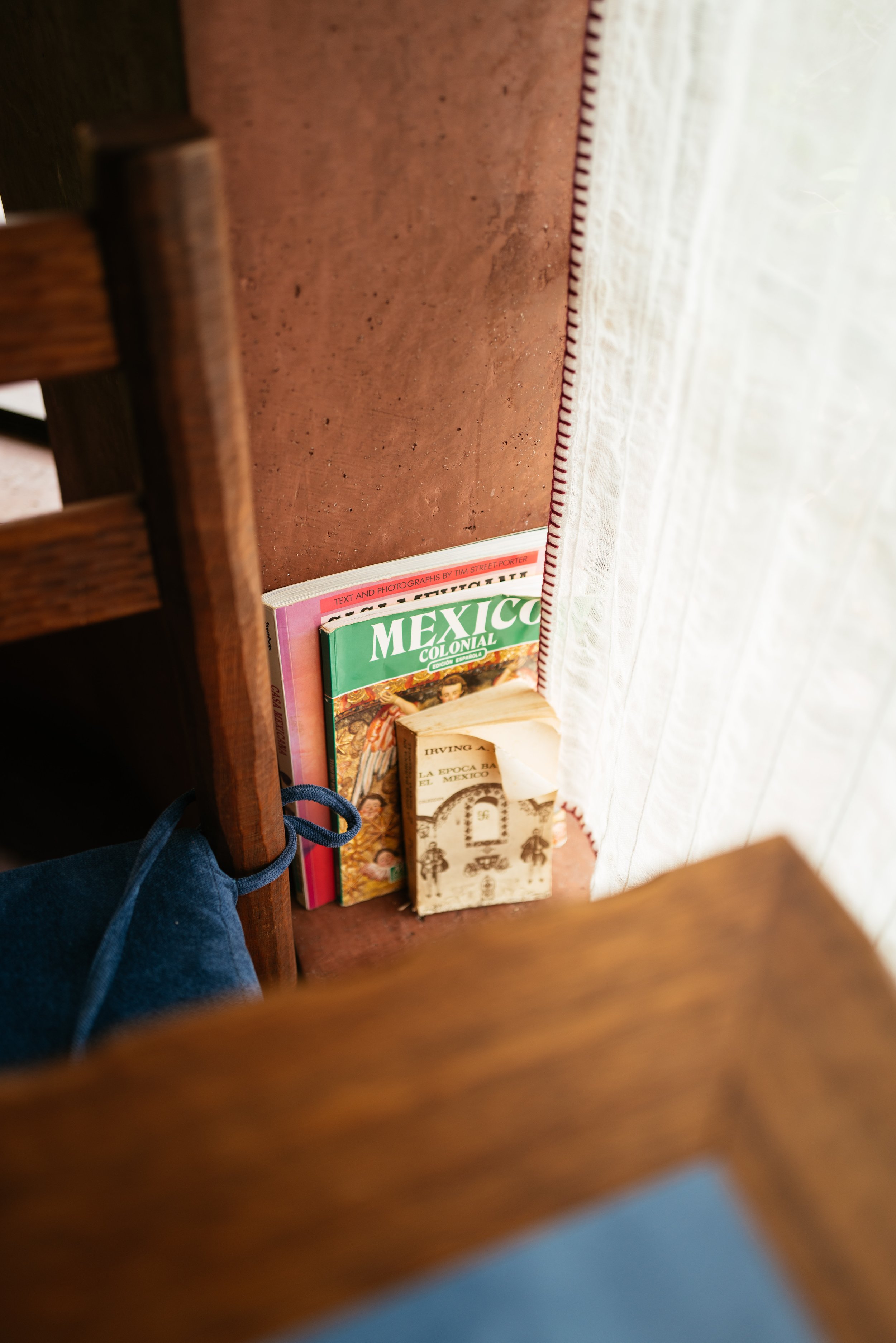 View from underneath a wooden chair looking at a small shelf with books, a book with a tan cover, a book with a green cover titled 'Mexico Colonial,' and another book with a pink cover. Part of a white curtain is visible on the right.