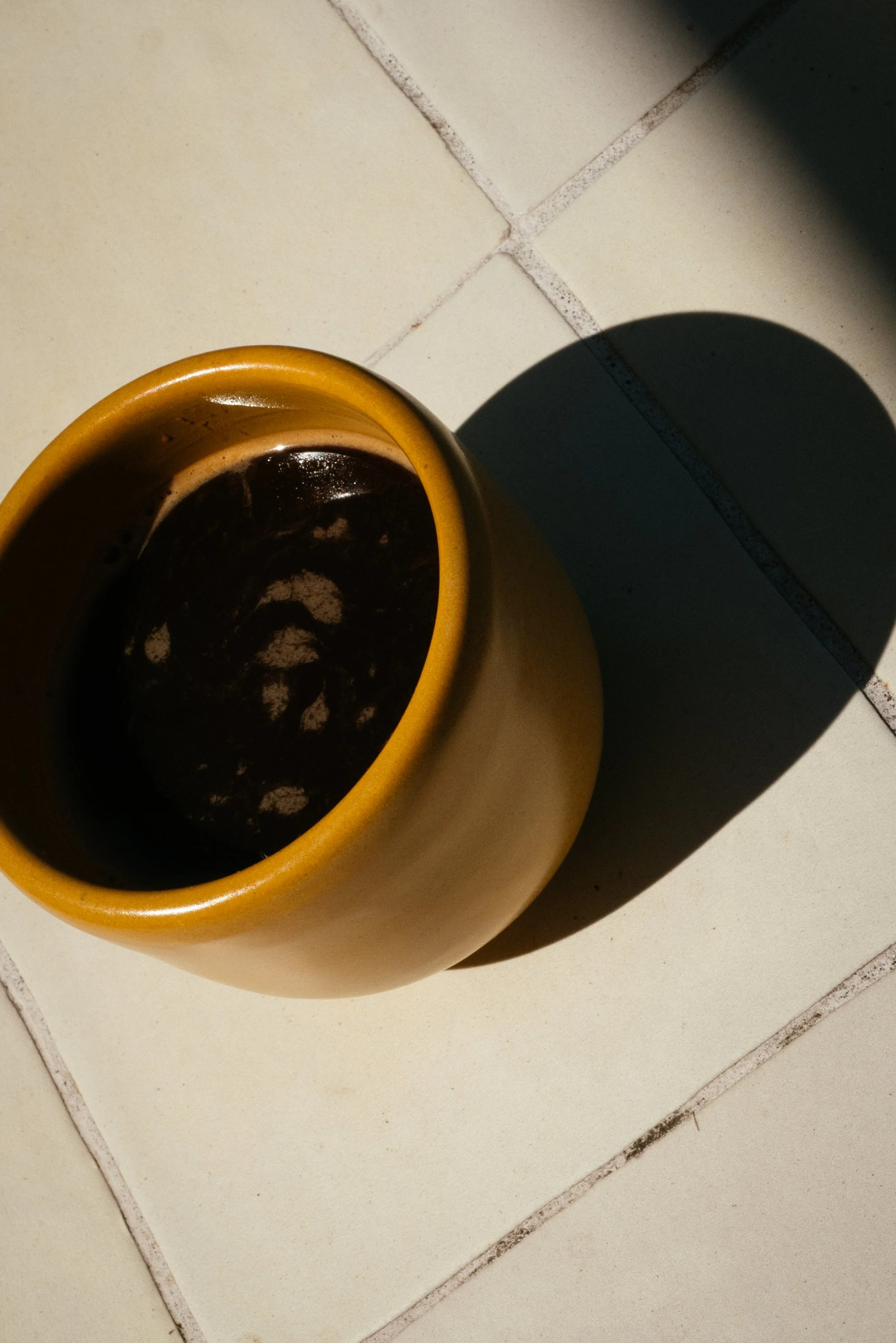 Yellow coffee mug on a tiled floor with black coffee inside, casting a shadow.