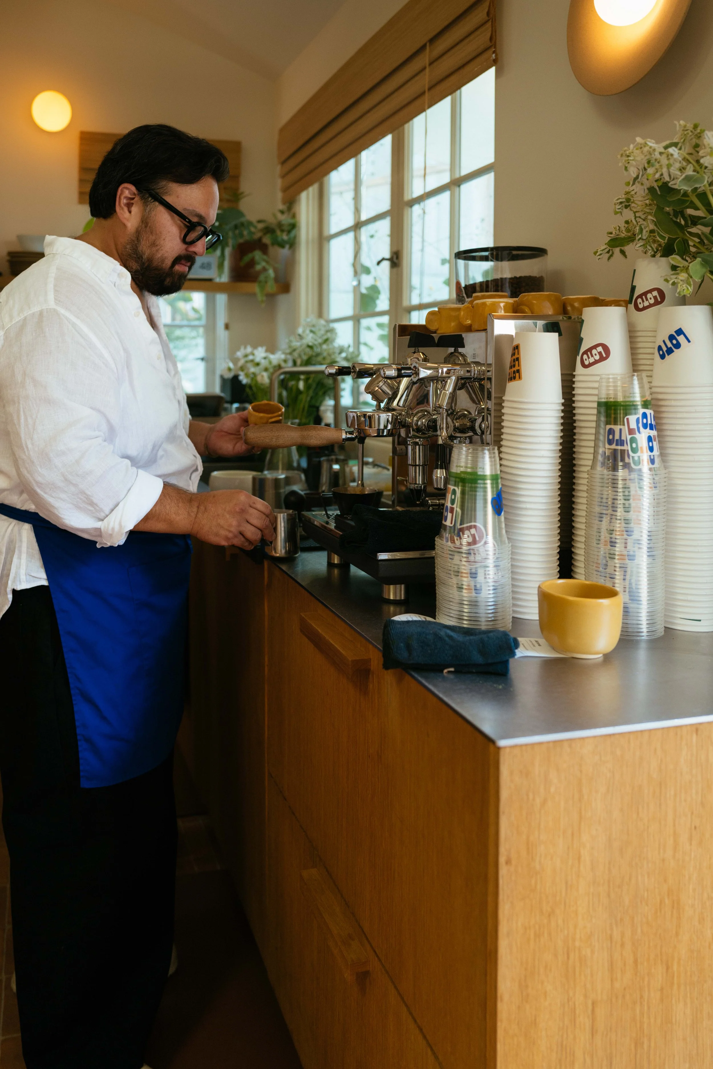 A man with dark hair, glasses, and a beard in a white shirt and blue apron makes coffee at a wooden counter in a cozy cafe. There are cups, a coffee grinder, plants, and flowers in the background with sunlight coming through large windows.