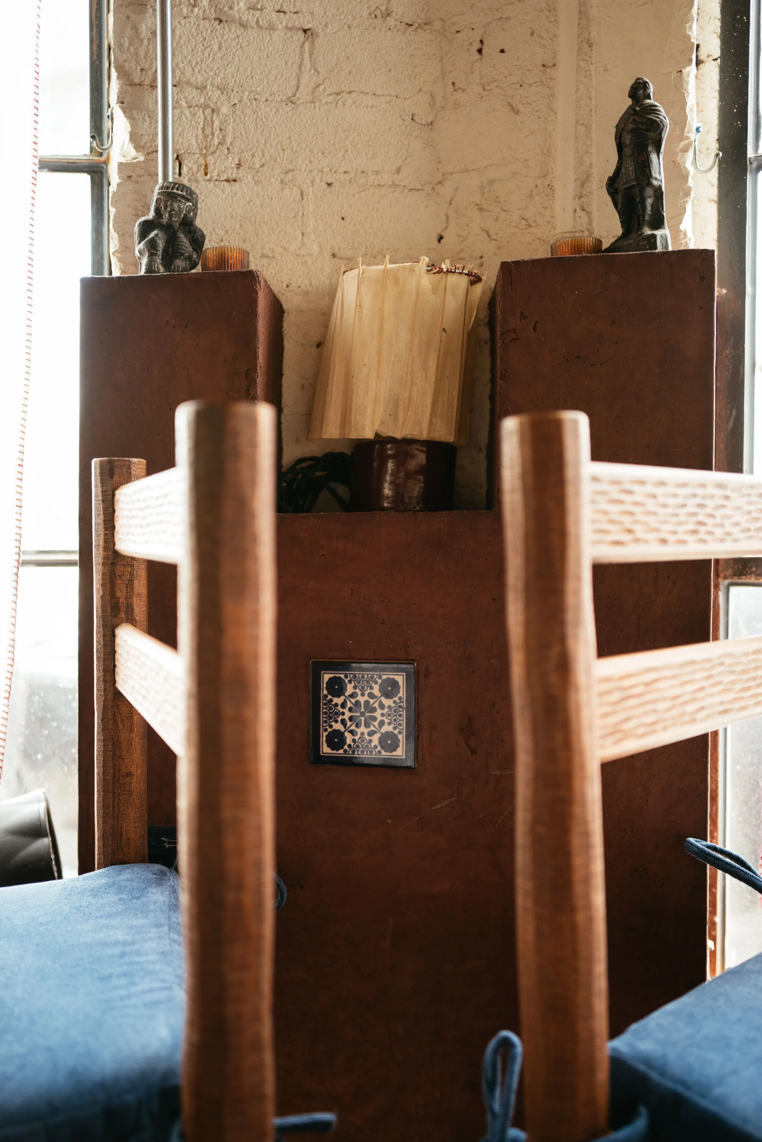 Interior view of a room with a rustic brick wall, two small statues on brick pedestals, a book, and wooden chairs.