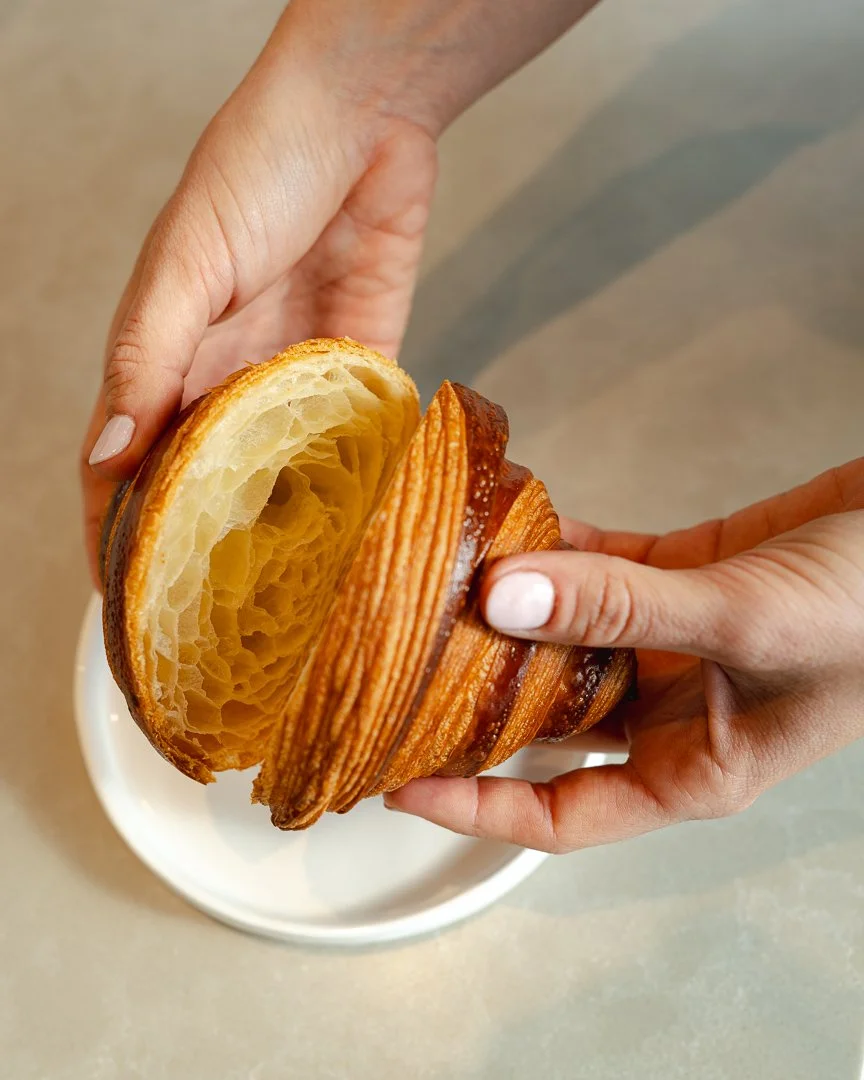 Hands pulling apart a flaky croissant to show its layers, Louma Bakery