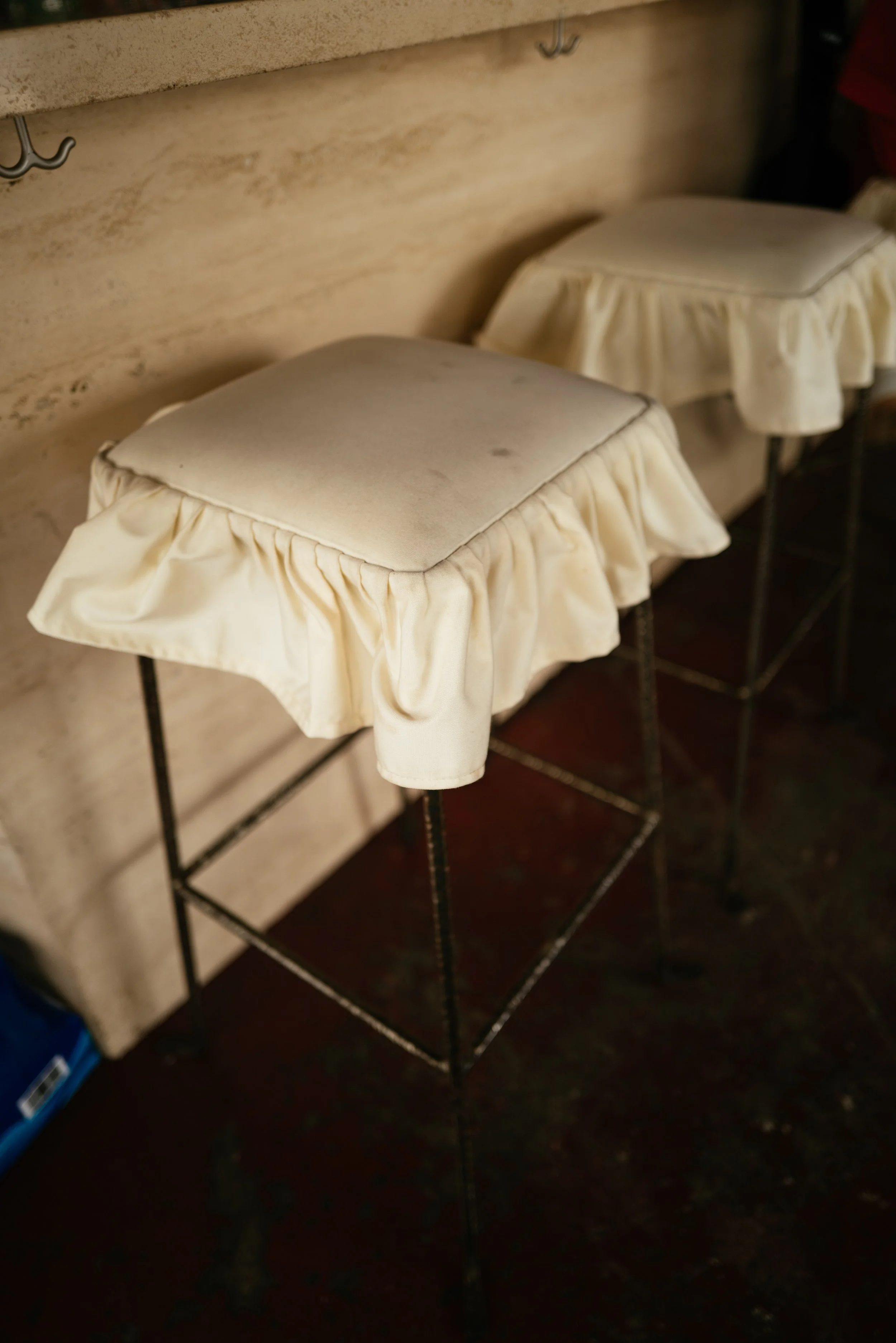 Two bar stools with cream-colored cushioned seats and ruffled skirts, placed against a beige wall with hooks.