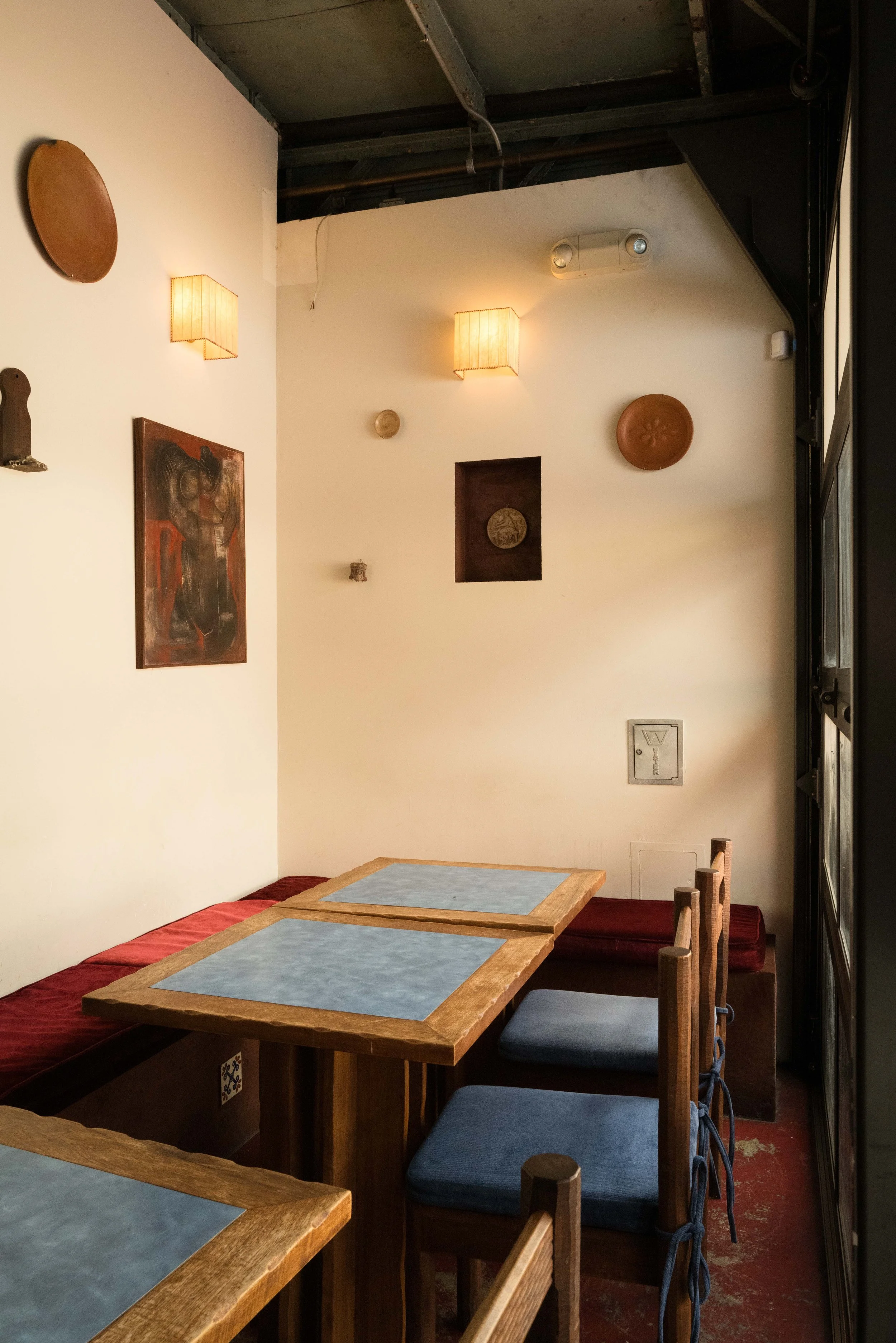 Interior of a cozy restaurant with wooden tables and chairs with blue cushions, white walls adorned with various decorative plates and artwork, warm lighting from wall sconces, and a partially visible window on the right side.