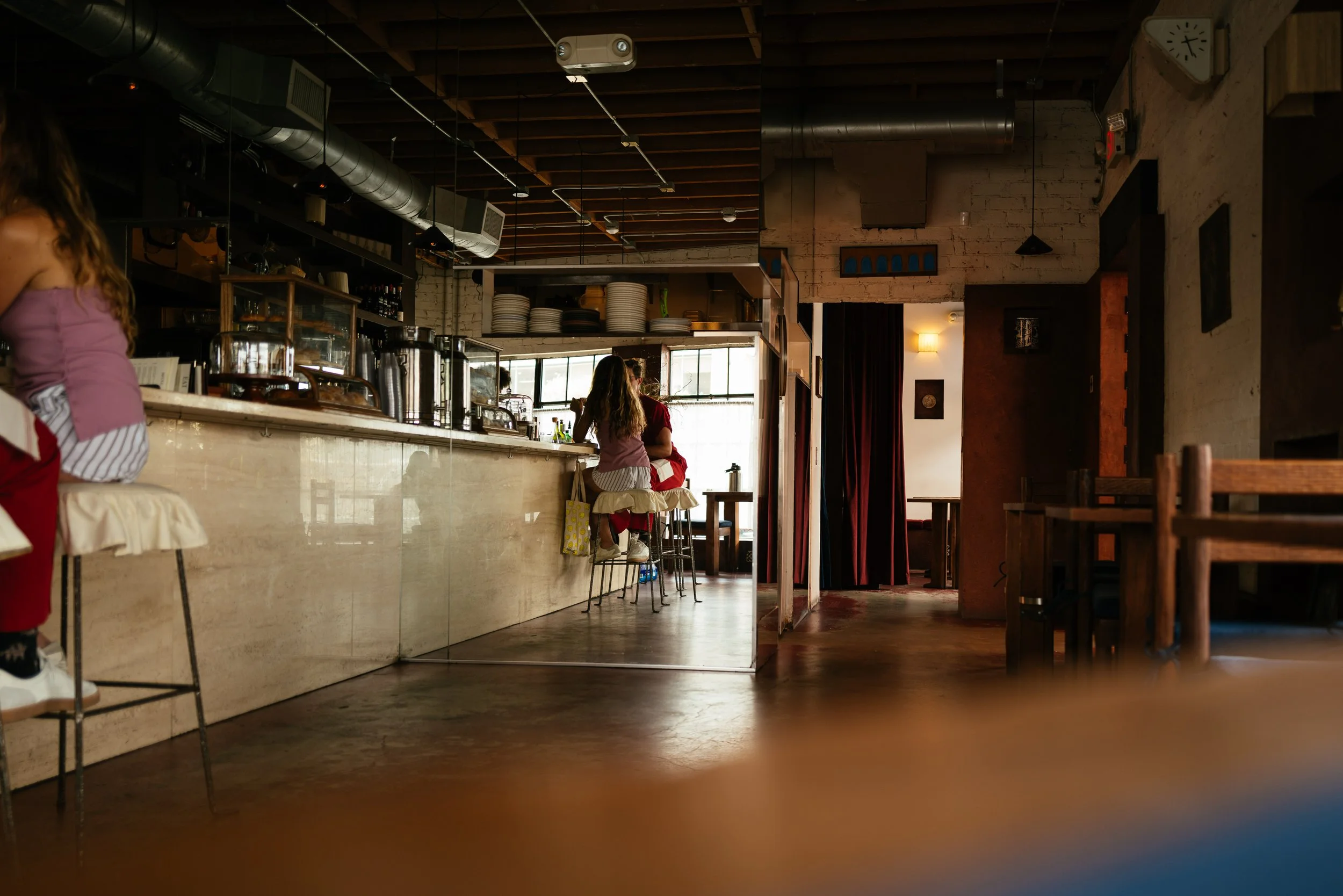 Interior of a modern restaurant or cafe with a counter, a couple of people sitting on bar stools, and a person partially visible on the left. The space features exposed brick walls, wooden beams, and industrial ductwork on the ceiling.