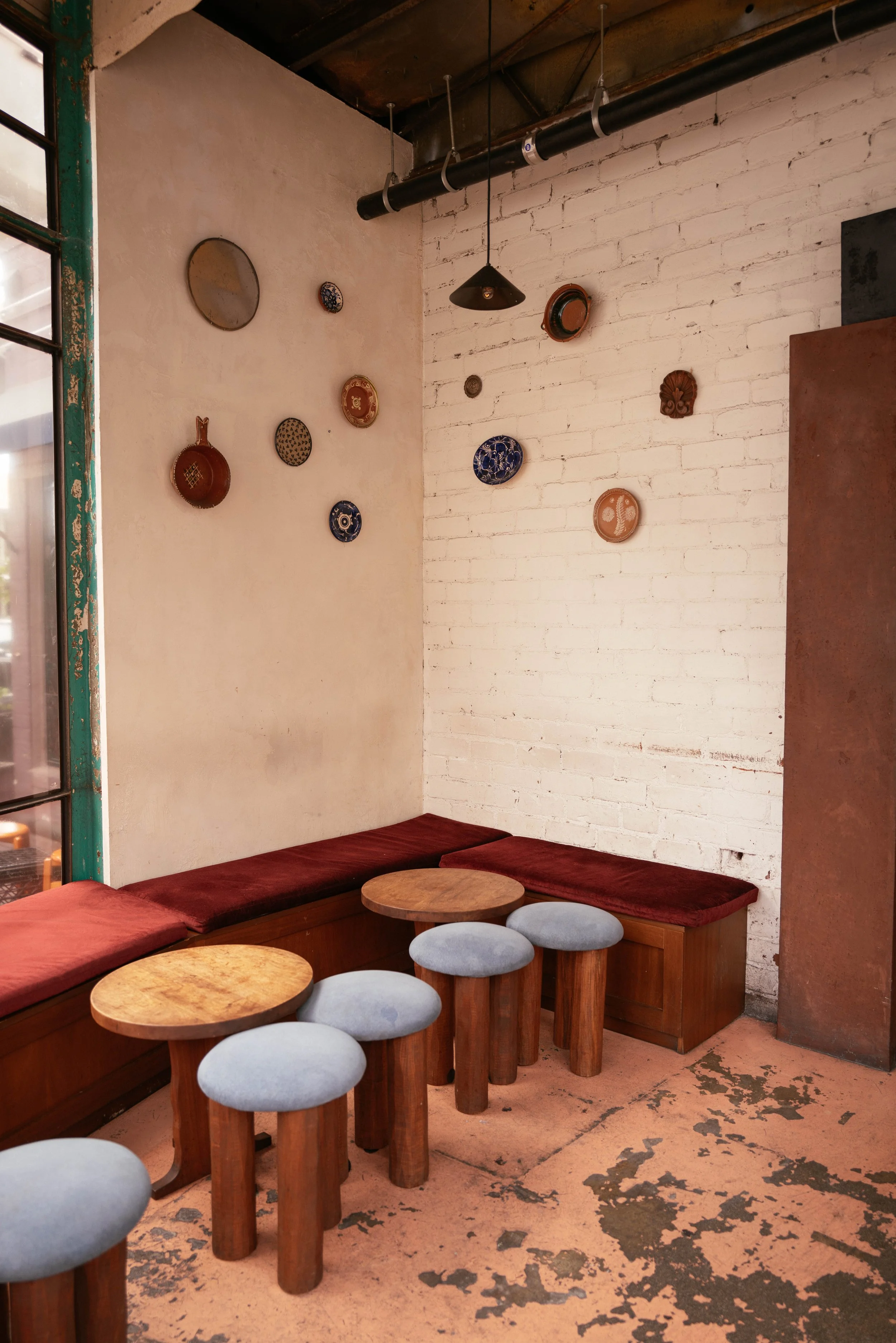 Interior of a cozy cafe with a white brick wall decorated with various plates, wooden tables, and padded stools in a corner with a built-in bench with red cushions.